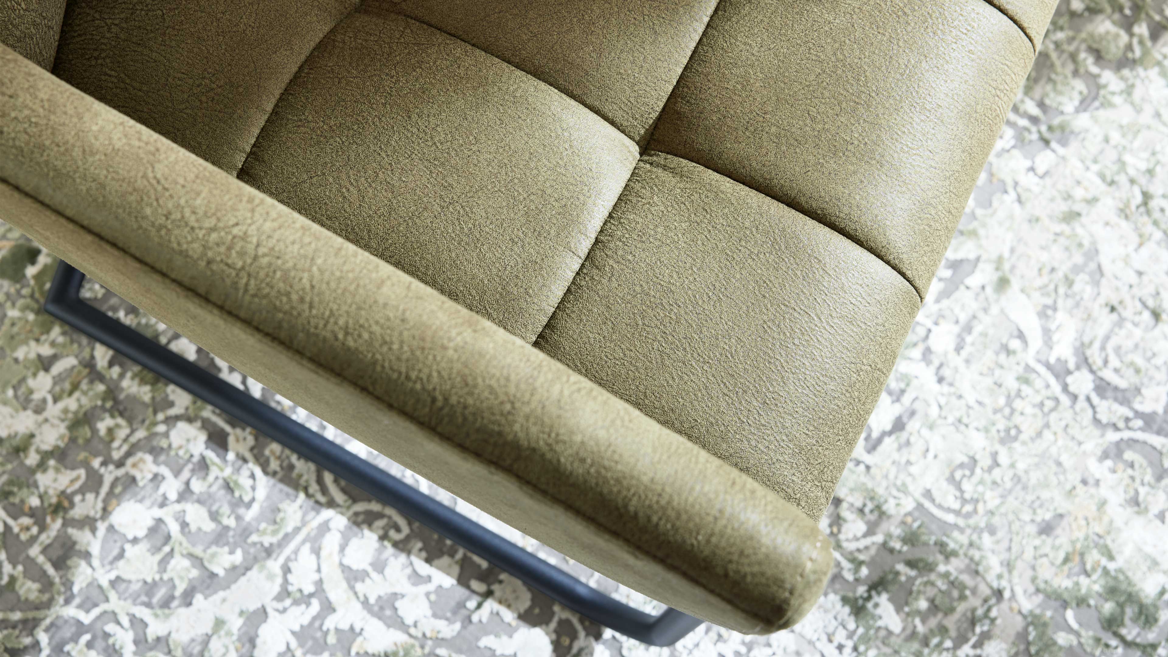 Olivegrün gepolsterter Stuhl mit genähter Karosteppung, abgerundetem Armteil und grauem Metallgestell auf floralem Teppich.