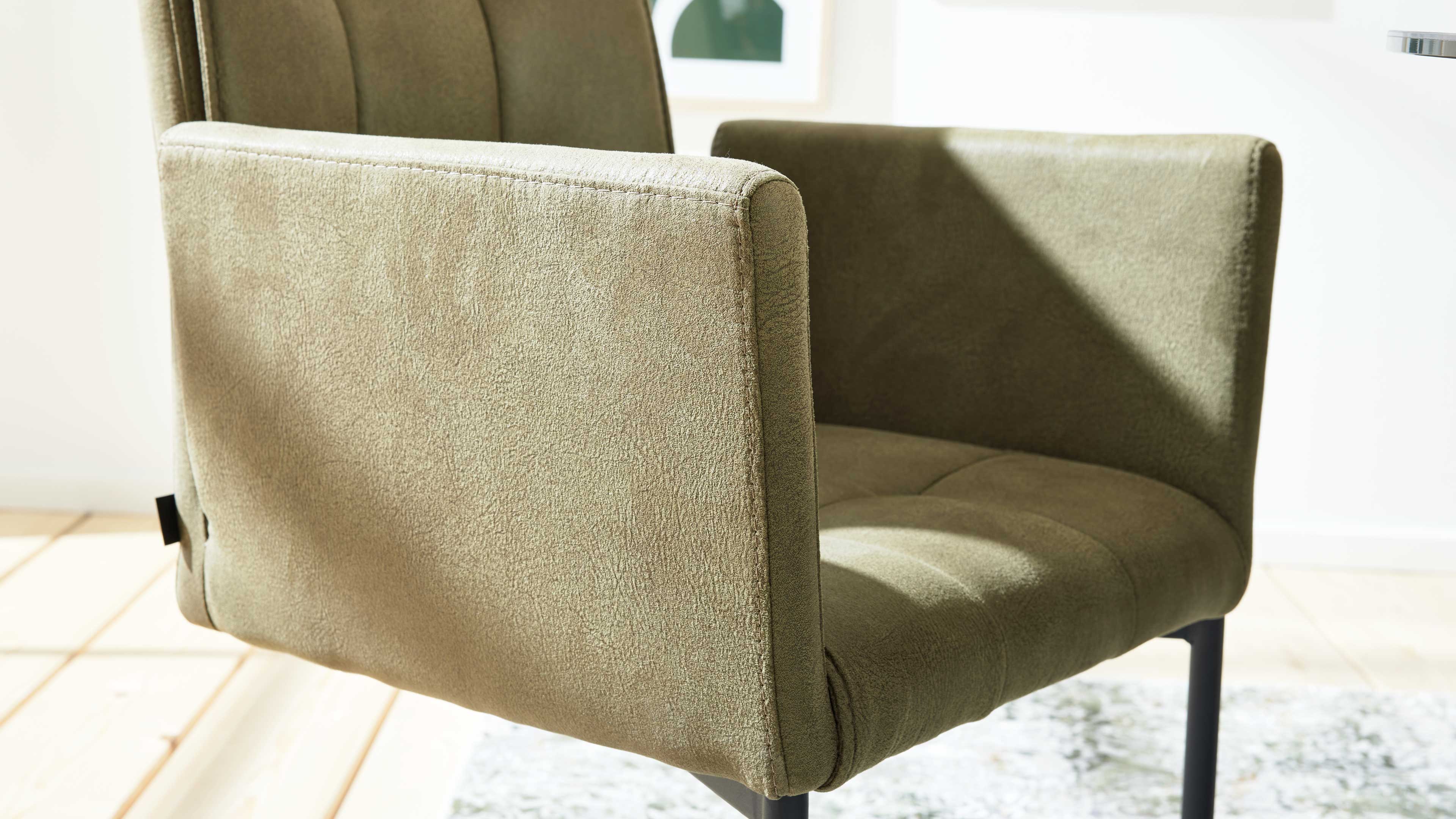 Nahaufnahme eines olivgrünen gepolsterten Sessels mit quadratischen Armlehnen, gesteppter Sitzfläche und schwarzen Beinen im Sonnenlicht.