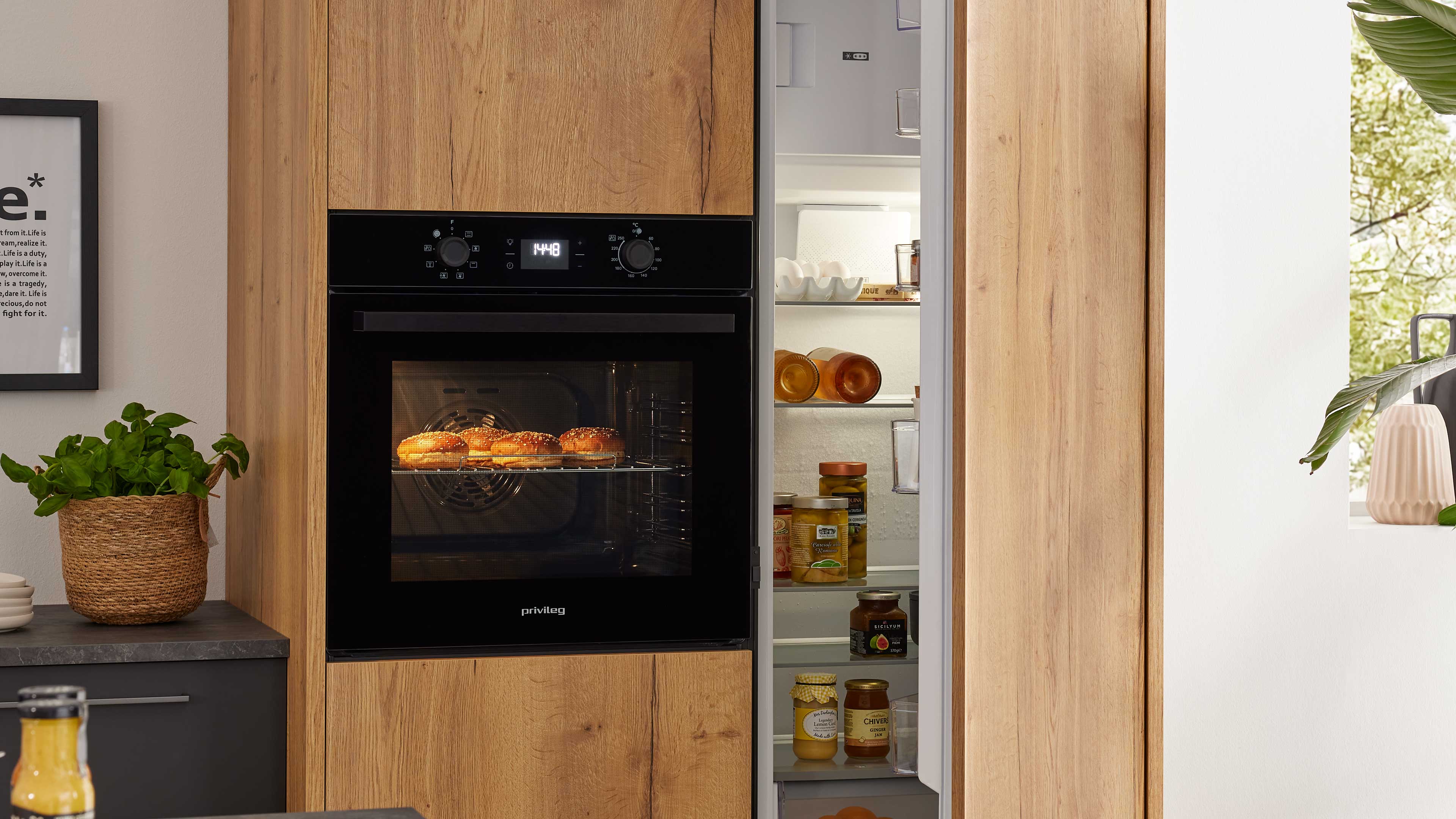Eingebauter schwarzer Backofen in Holzschrank mit gebackenen Brötchen im Inneren und geöffnetem Kühlschrank daneben.
