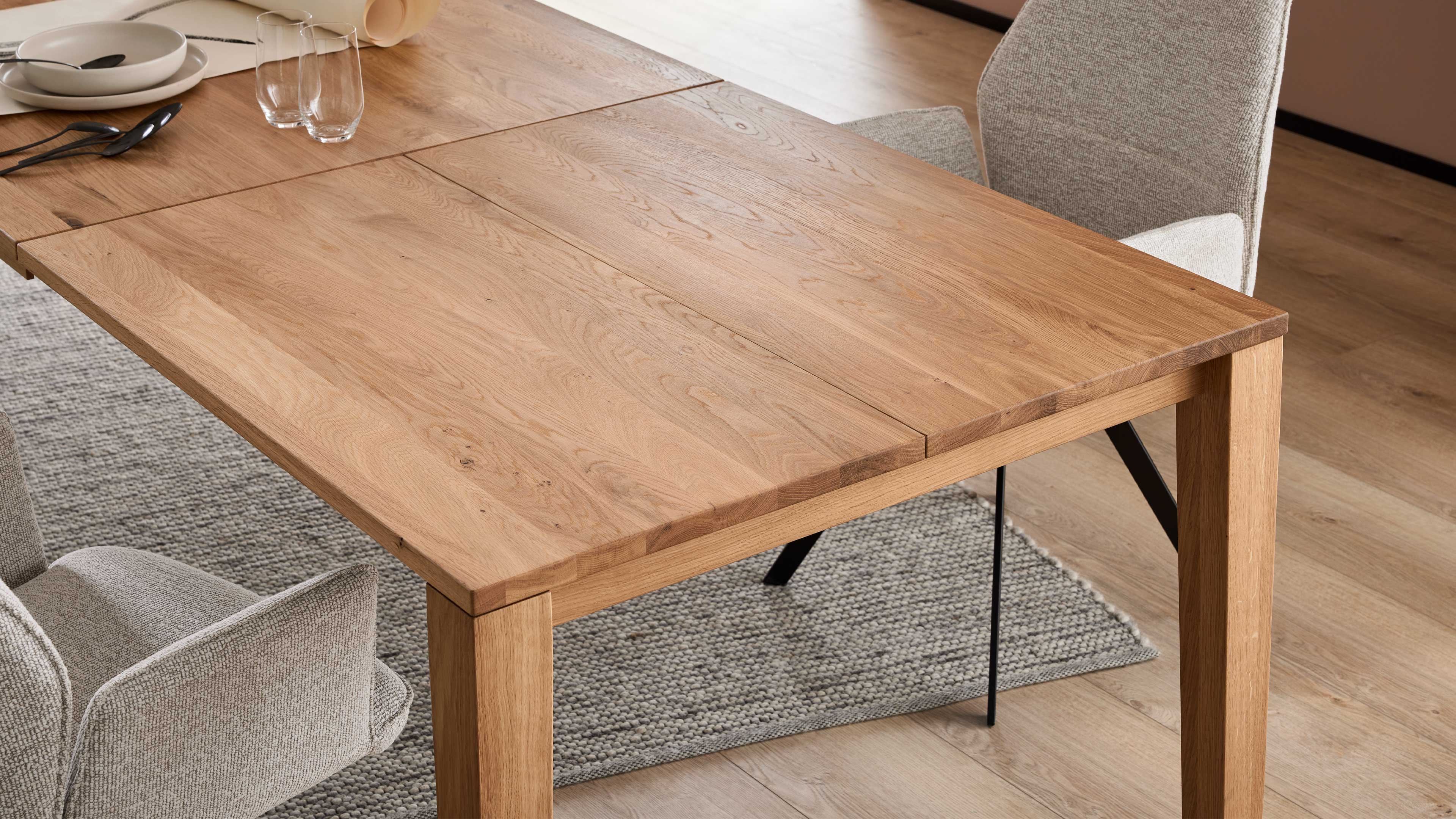 Massiver rechteckiger Esstisch aus hellem Holz mit sichtbarer Maserung, zwei gepolsterte Stoffstühle, Teppich und Tischgeschirr.