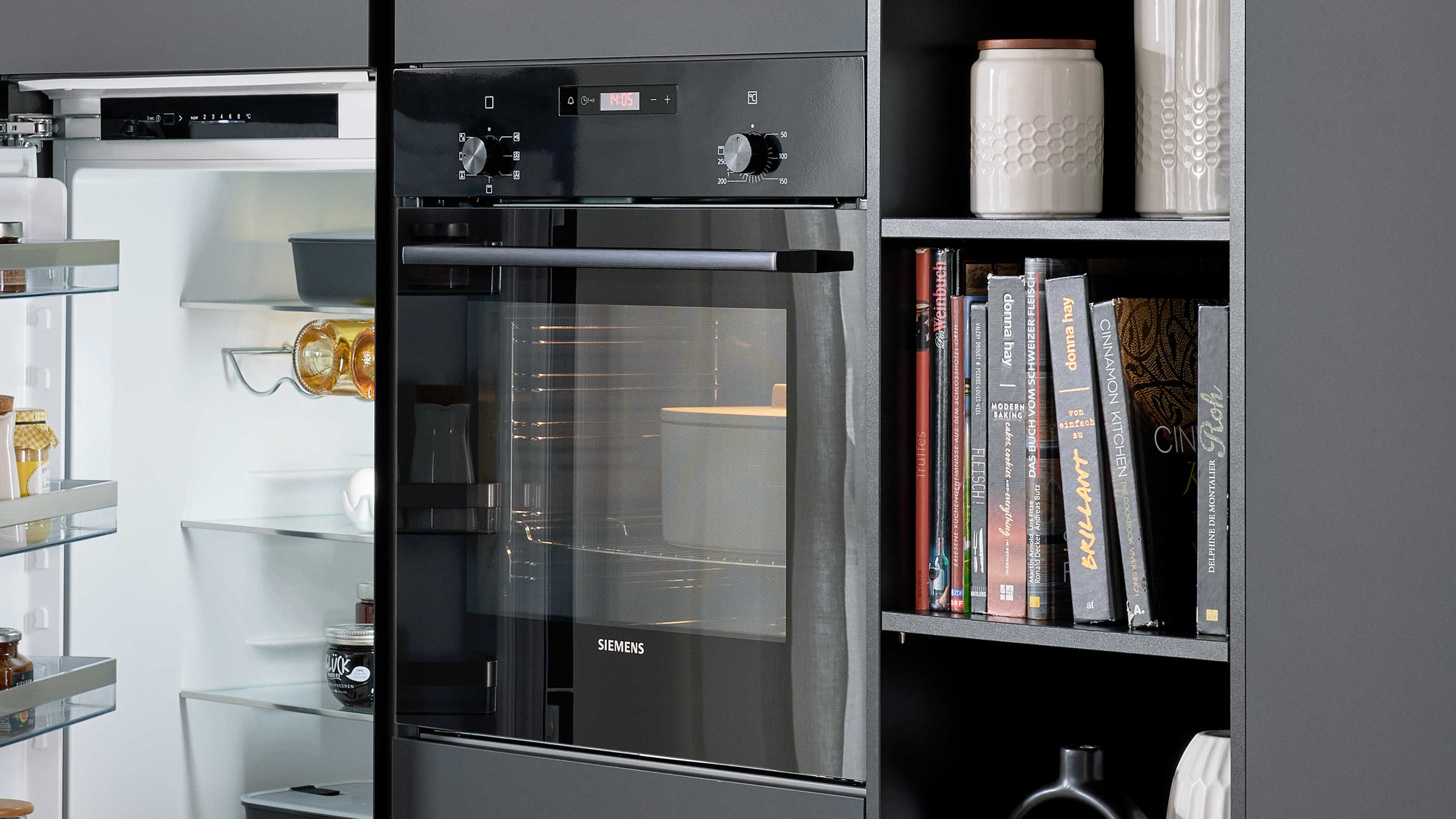 Schwarzer Siemens Einbaubackofen mit Glasfront zwischen offenem Kühlschrank links und offenem Regal mit Gläsern, Büchern und Keramik rechts.