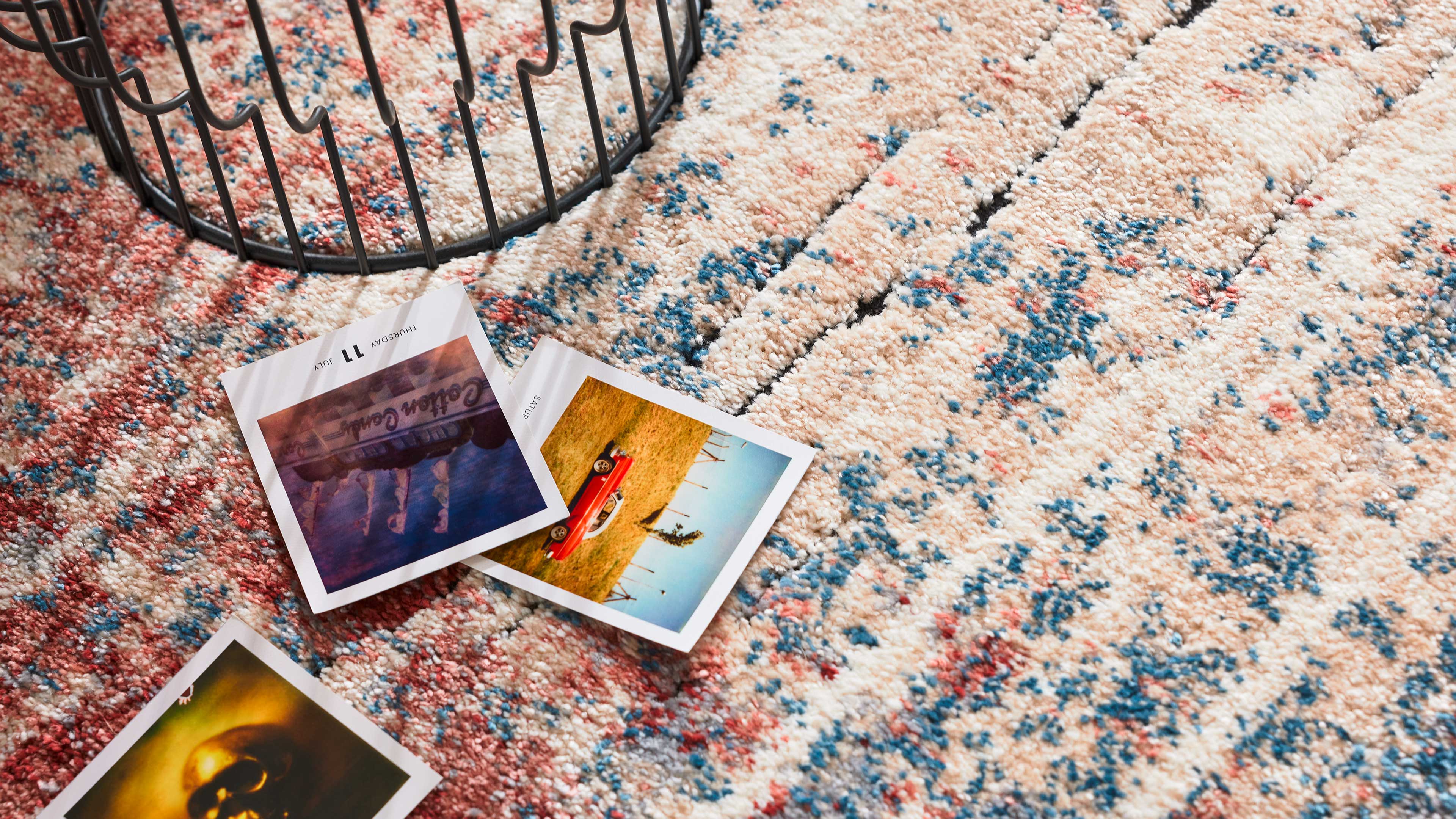 Beiger hochfloriger Teppich mit verstreuten blauen und roten Sprenkeln, darauf drei Polaroid-Fotos und der Rand eines schwarzen Drahtkorbs.