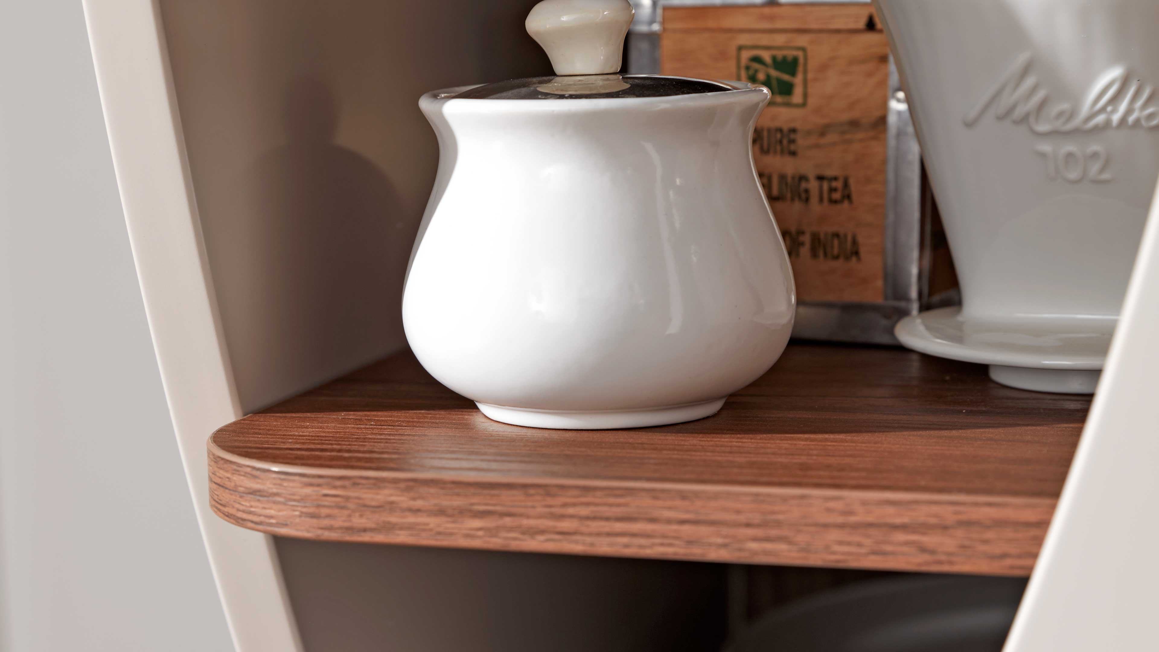 Auf einer hellbraunen Holzablage steht ein bauchiges weißes Keramikgefäß mit Deckel, daneben ein weißes Keramikteil und eine Holzbox.