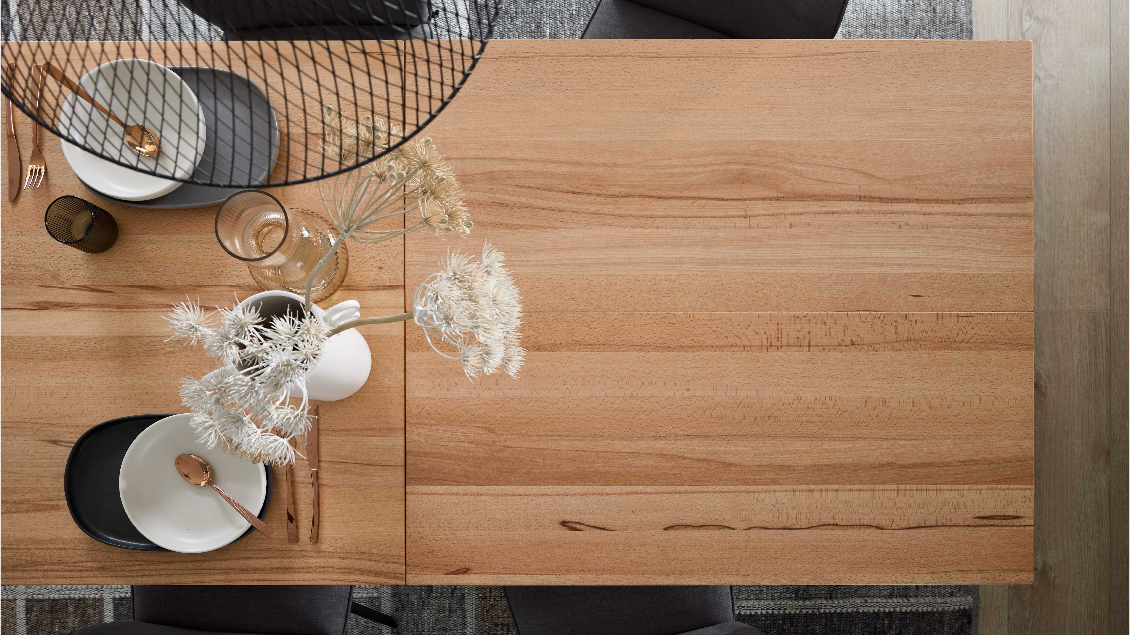 Rechteckige helle Holz-Tischplatte mit Gedeck, goldenem Besteck, Glas, weißer Vase mit getrockneten Blüten und Drahtlampenschirm-Schatten.