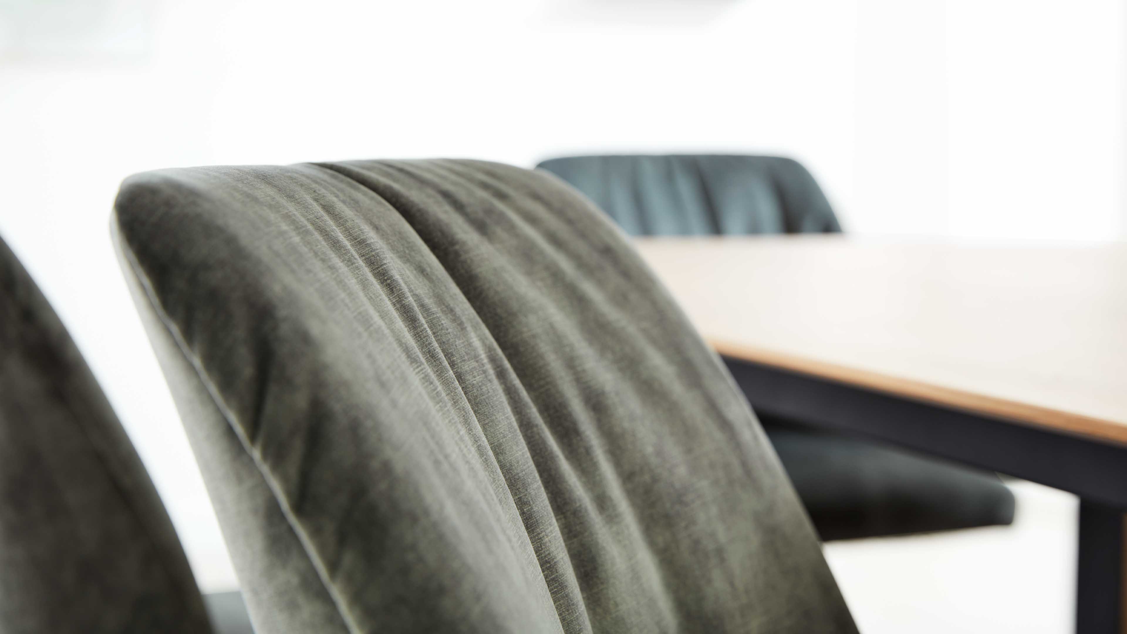 Nahaufnahme einer graugrünen gepolsterten Stuhllehne mit senkrechten Nähten, im Hintergrund ein Holztisch und weitere Stühle.