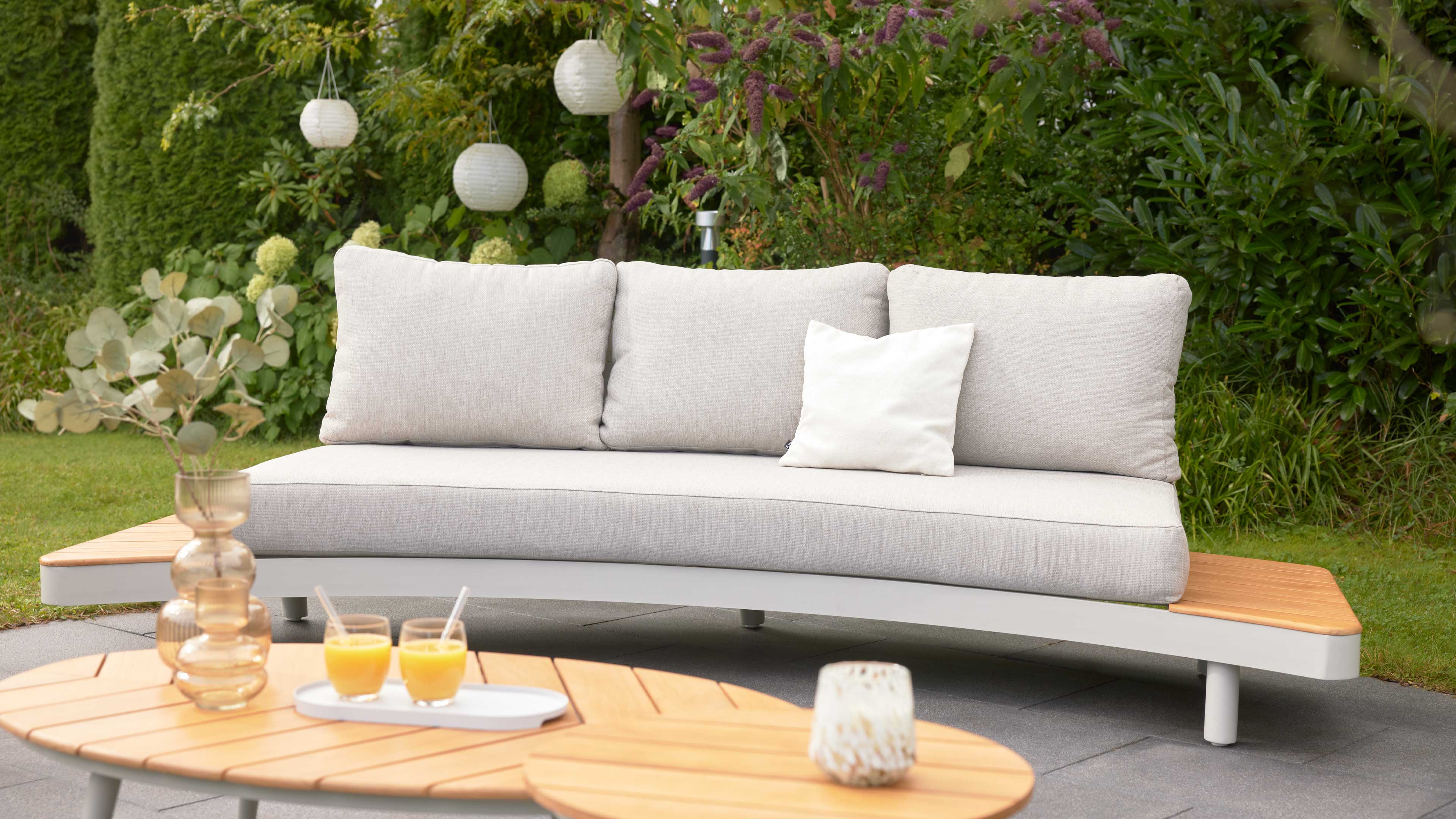 Geschwungene hellgraue Outdoor‑Sofa mit drei Rückenpolstern und kleinem weißen Kissen, Holzablagen an beiden Enden, vor grüner Hecke.