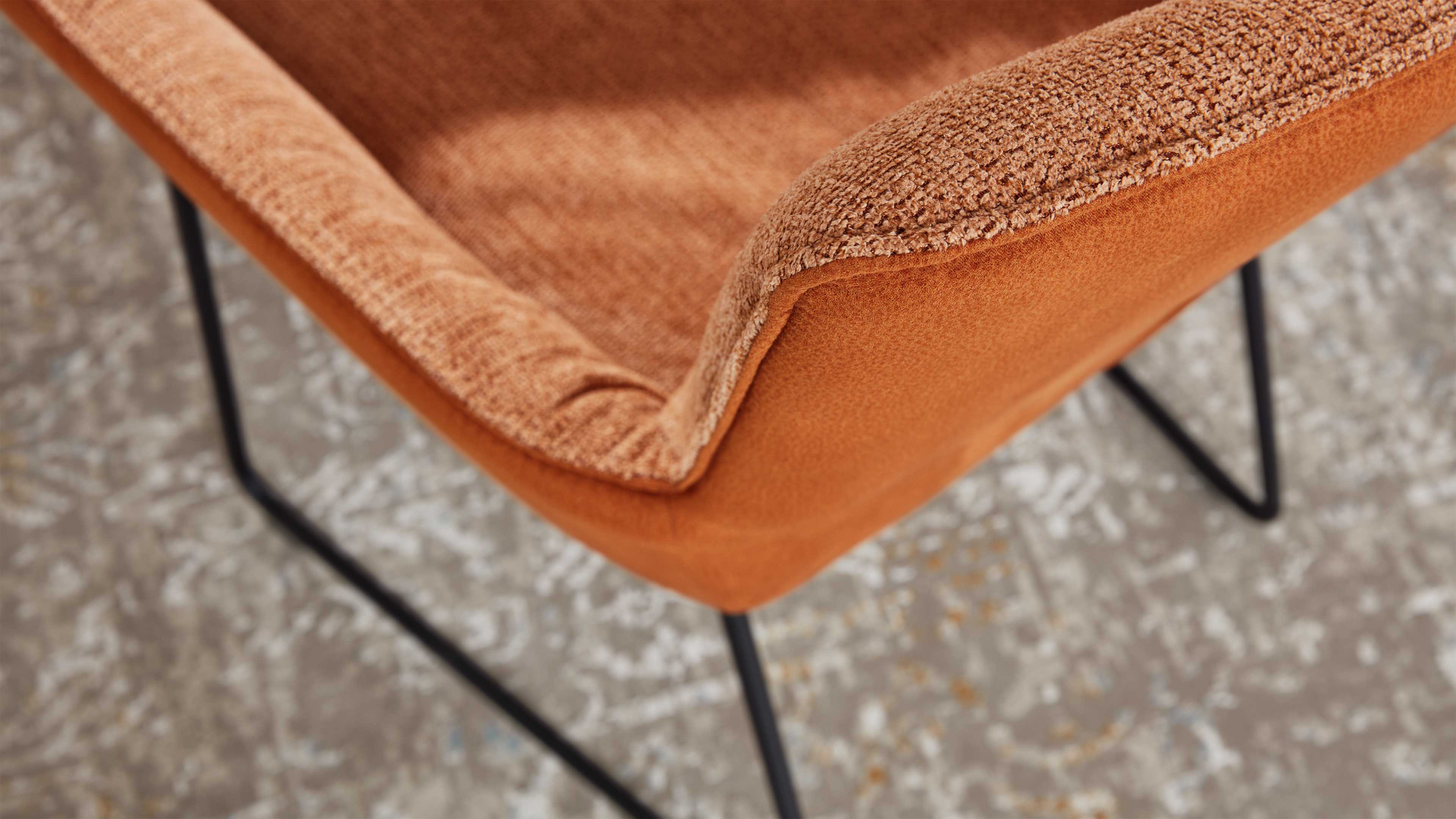 Nahaufnahme einer orangefarbenen, strukturierten Polsterung mit sichtbarer Naht, schwarzem Metallgestell und gemustertem Teppich.
