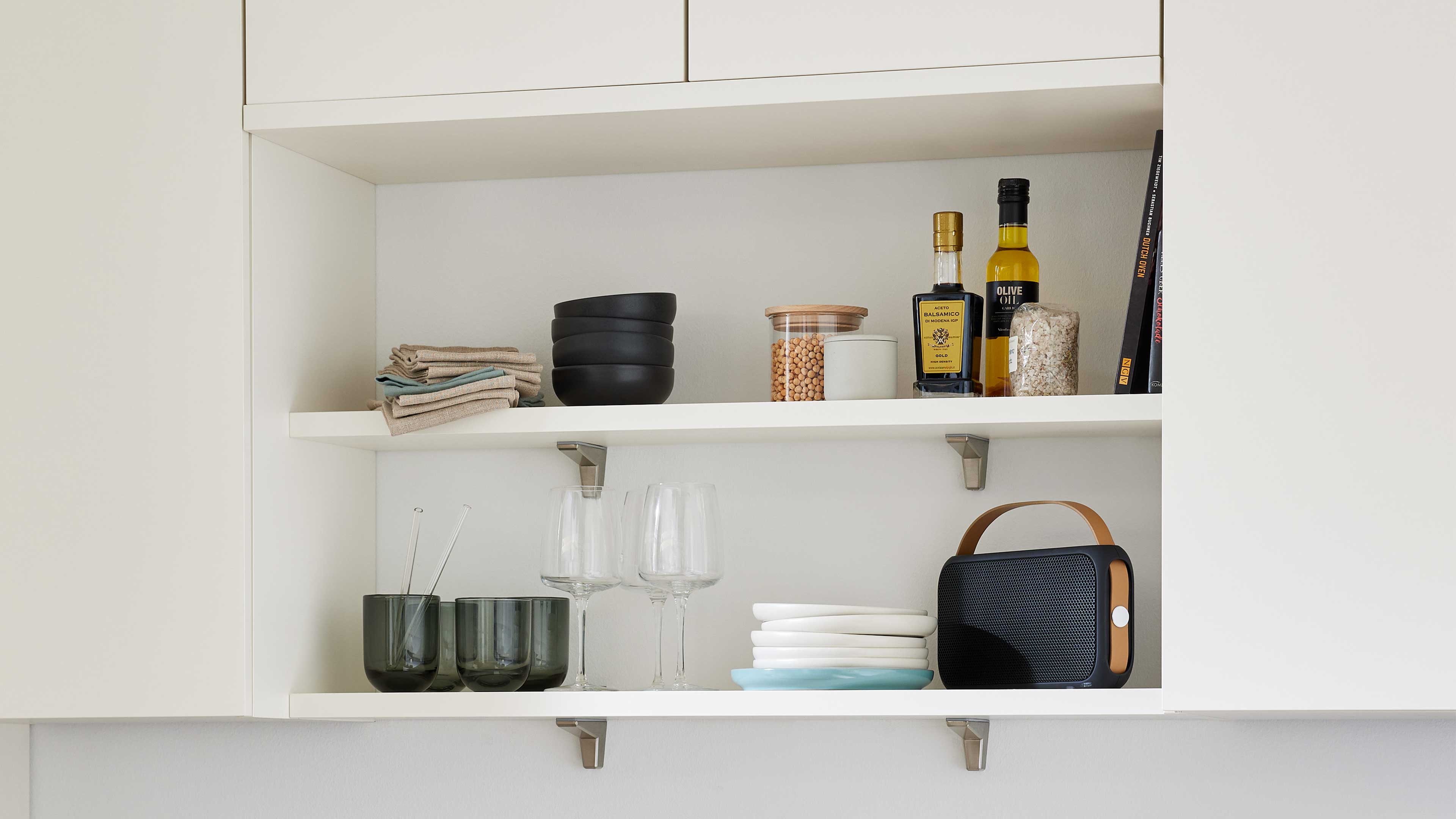 Offener weißer Küchenschrank mit zwei Regalböden, bestückt mit Schalen, Gläsern, Flaschen, Tellern und einem Lautsprecher.