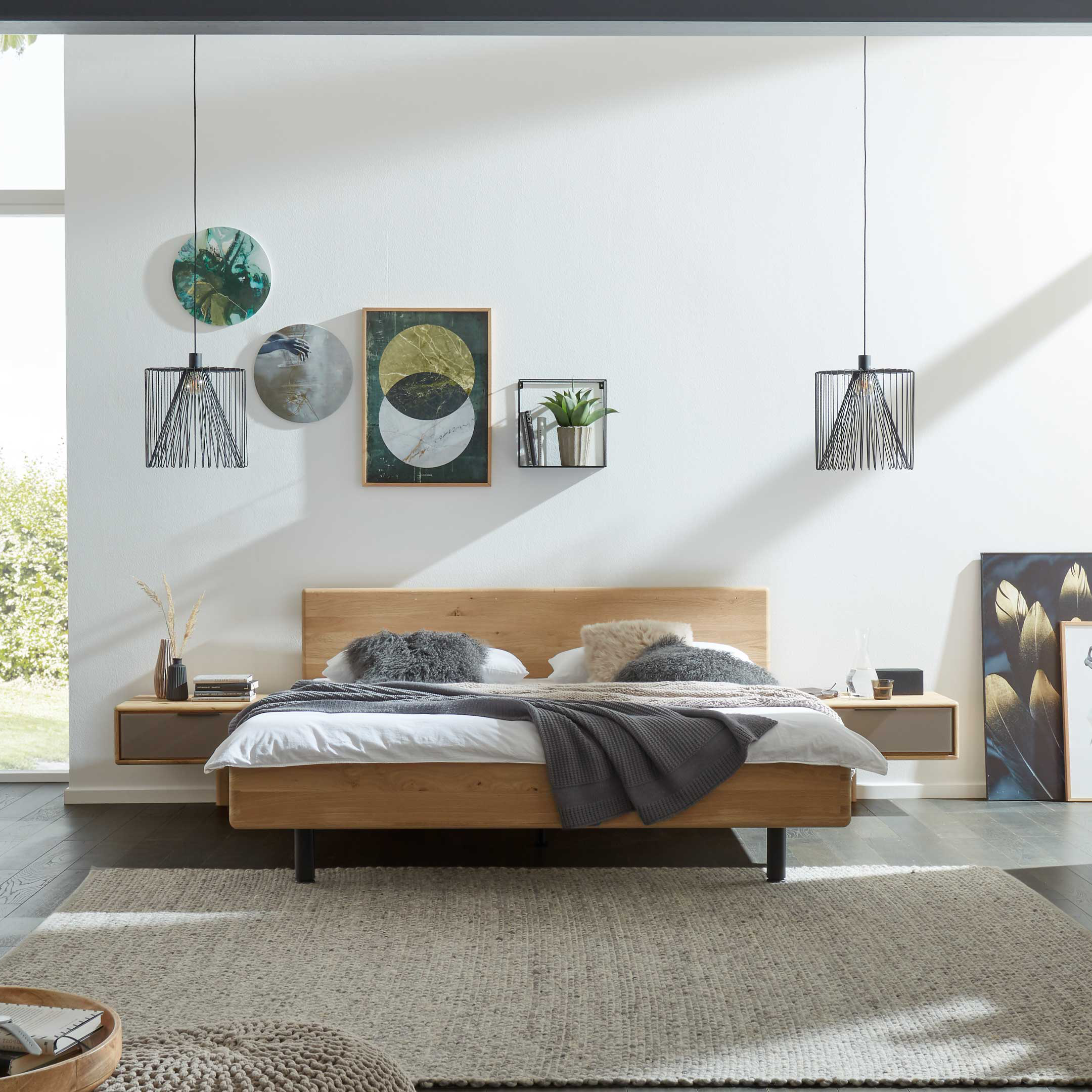 Holzbett mit Kopfteil, weißer Bettwäsche und grauer Decke, zwei hängende Drahtlampen, links ein Kleiderschrank und Fenster mit Gartenblick.