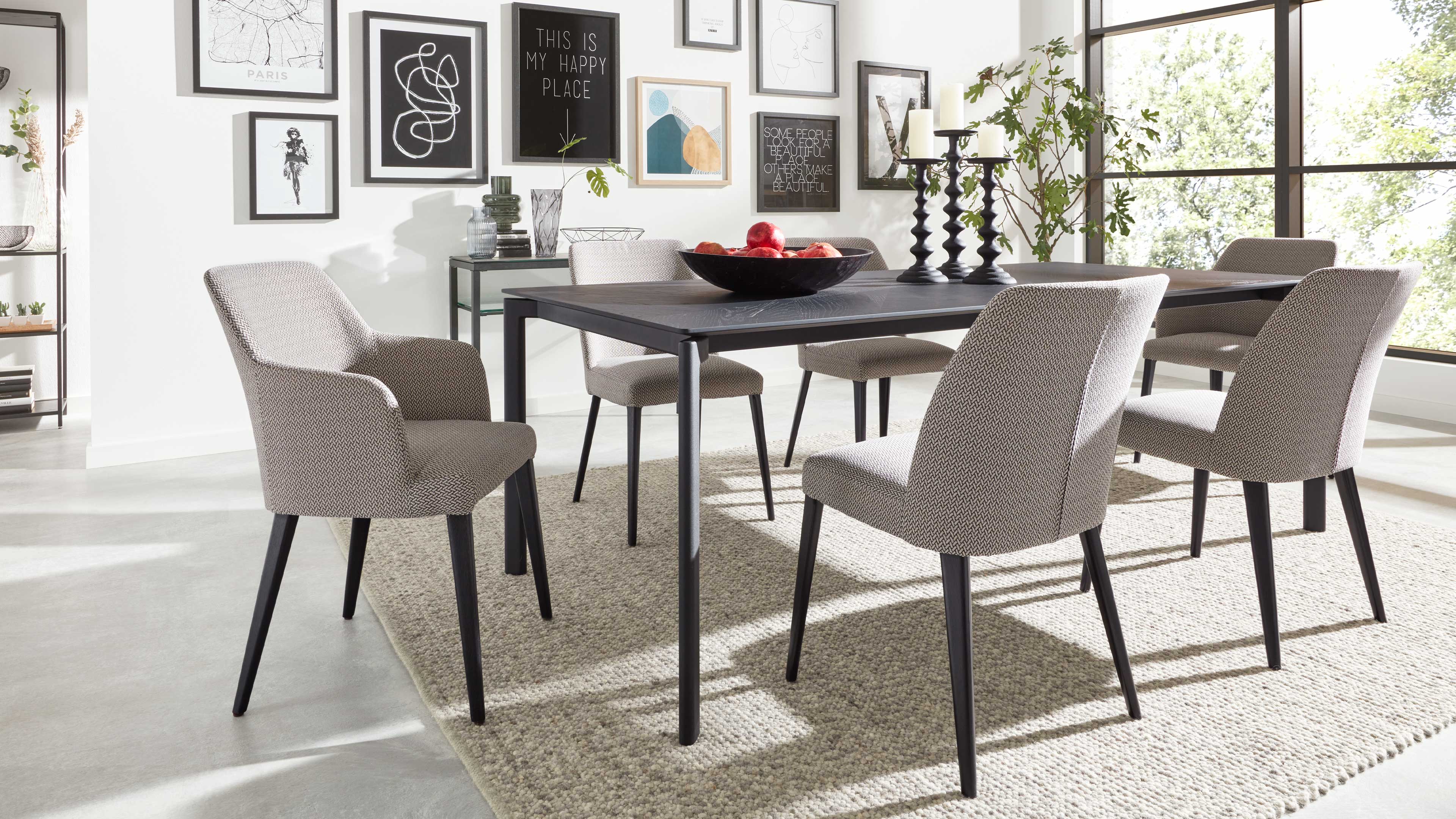 Schwarzer rechteckiger Esstisch auf hellem Teppich mit Schale voller Äpfel, Kerzenhaltern und sechs hellbezogenen Stühlen.