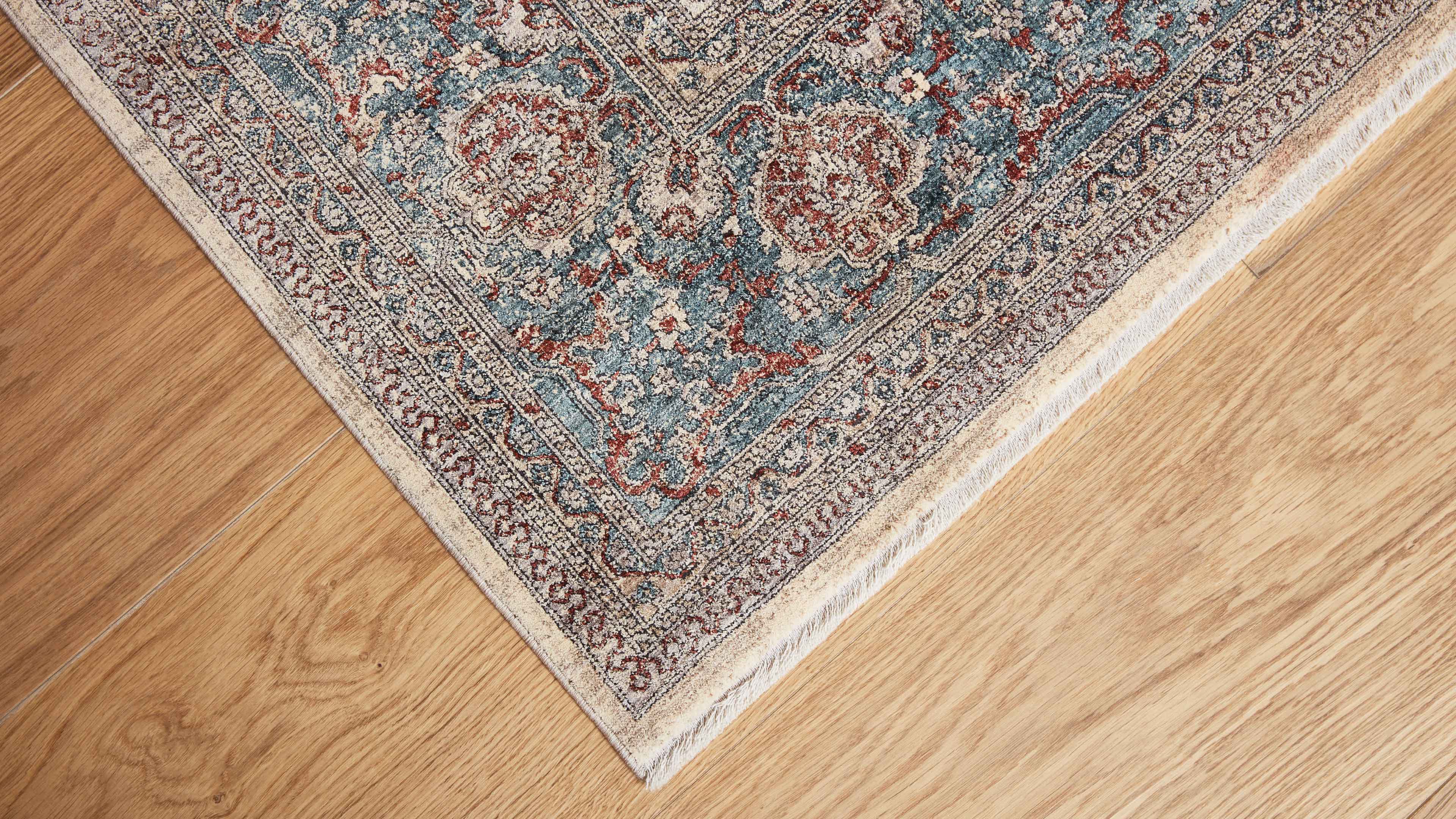 Teppickecke mit floralem Muster in Blau, Rot und Beige, dekorativem Bordürstreifen und kurzen weißen Fransen auf hellem Holzfußboden.