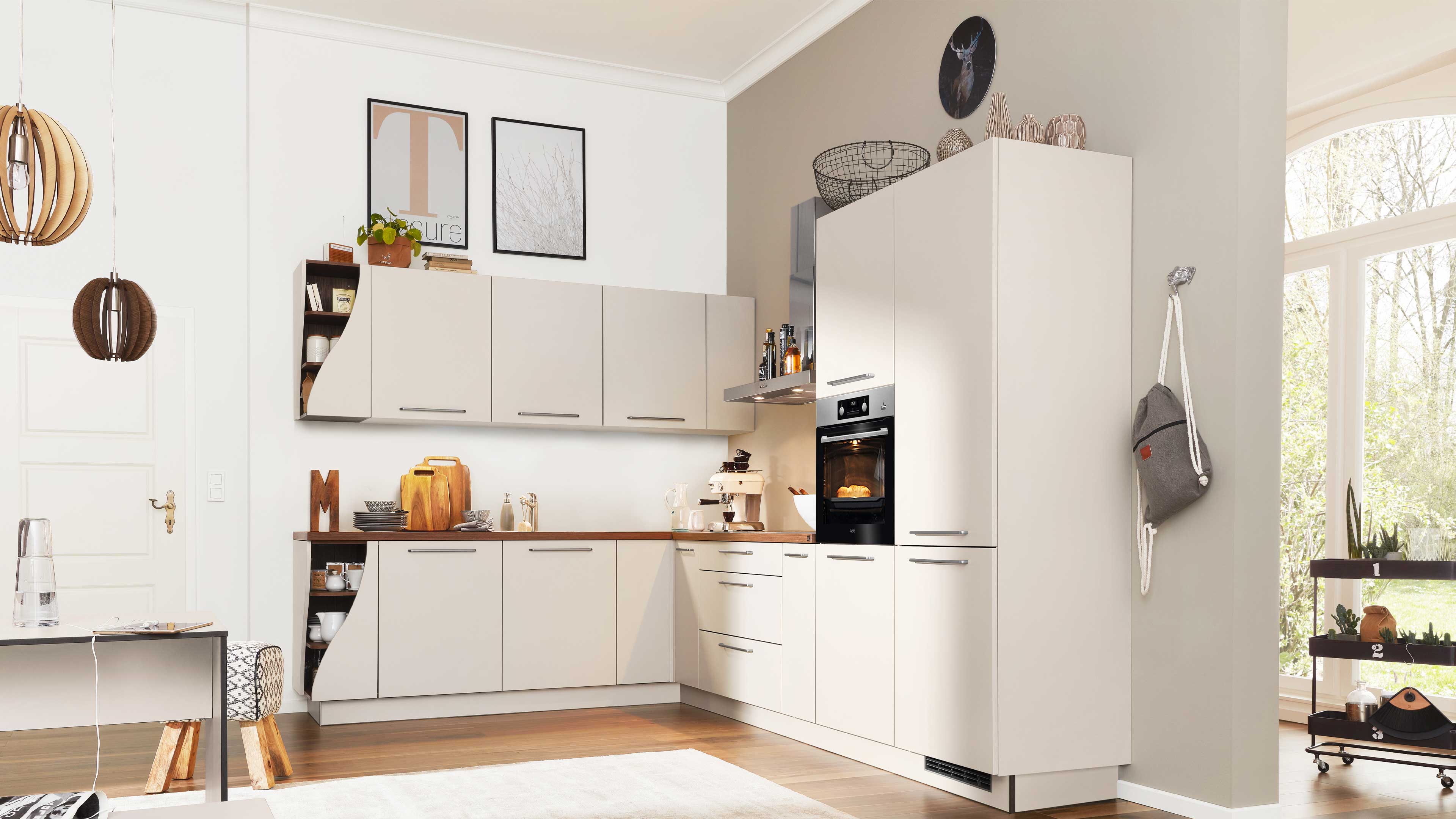 Beige L‑förmige Küchenzeile mit Holzarbeitsplatte, hohen Einbauschrank mit Backofen, flachen Fronten und Metallgriffen.