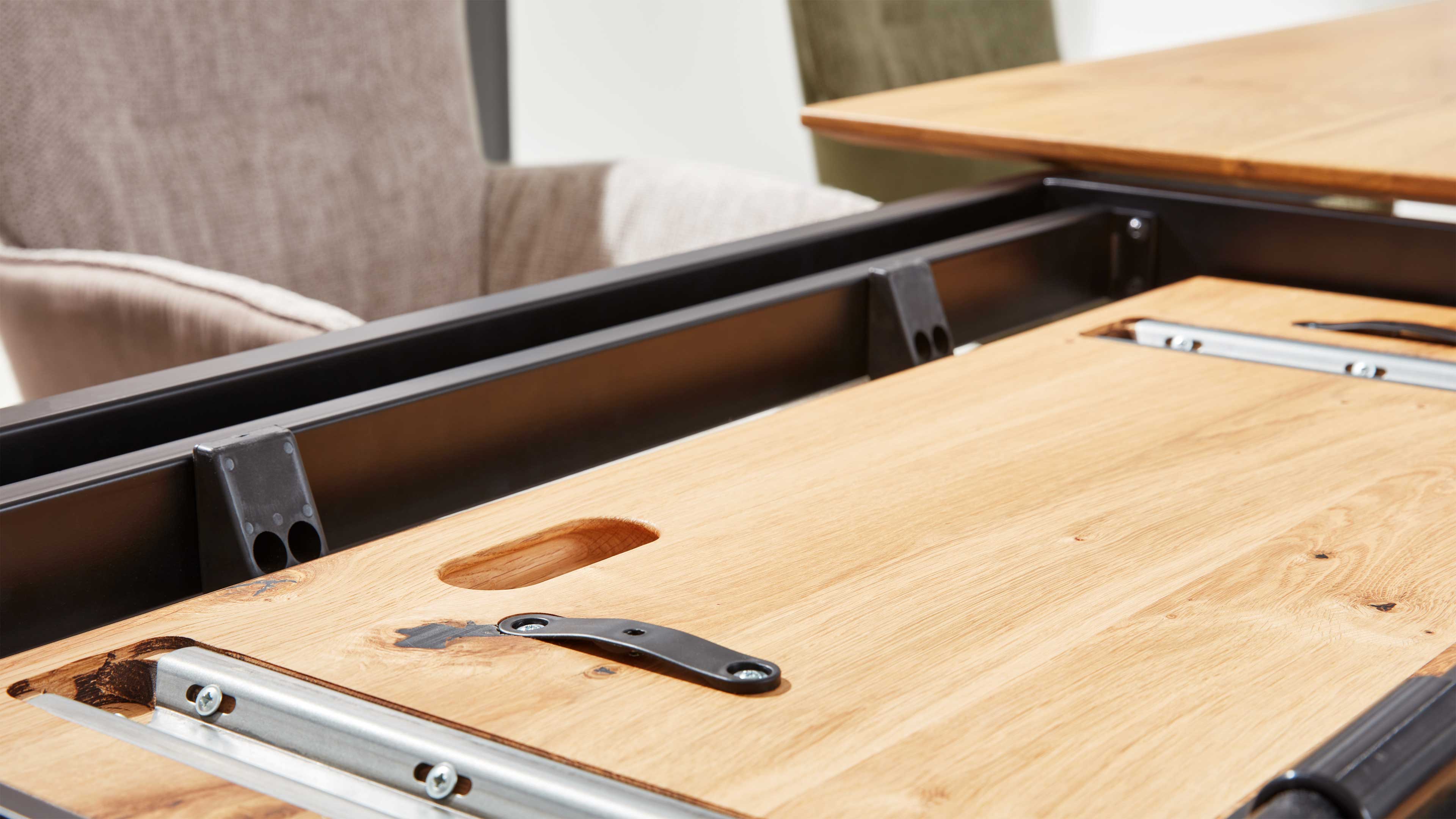 Ausgeklappte Holz-Tischplatte mit eingelassenem Griff, Metall-Schienen und schwarzem Gestell, dahinter ein beige gepolsterter Stuhl.