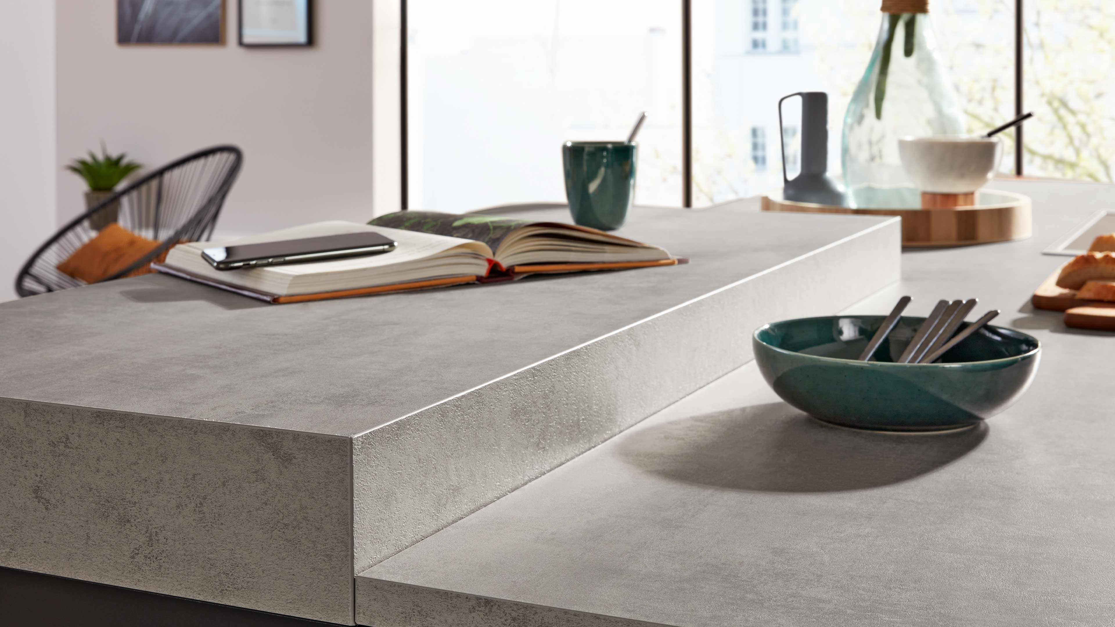 Zweistufige graue Küchenarbeitsplatte mit Beton-Optik, vorne grüne Schale mit Besteck, hinten offenes Buch, Smartphone und Becher.
