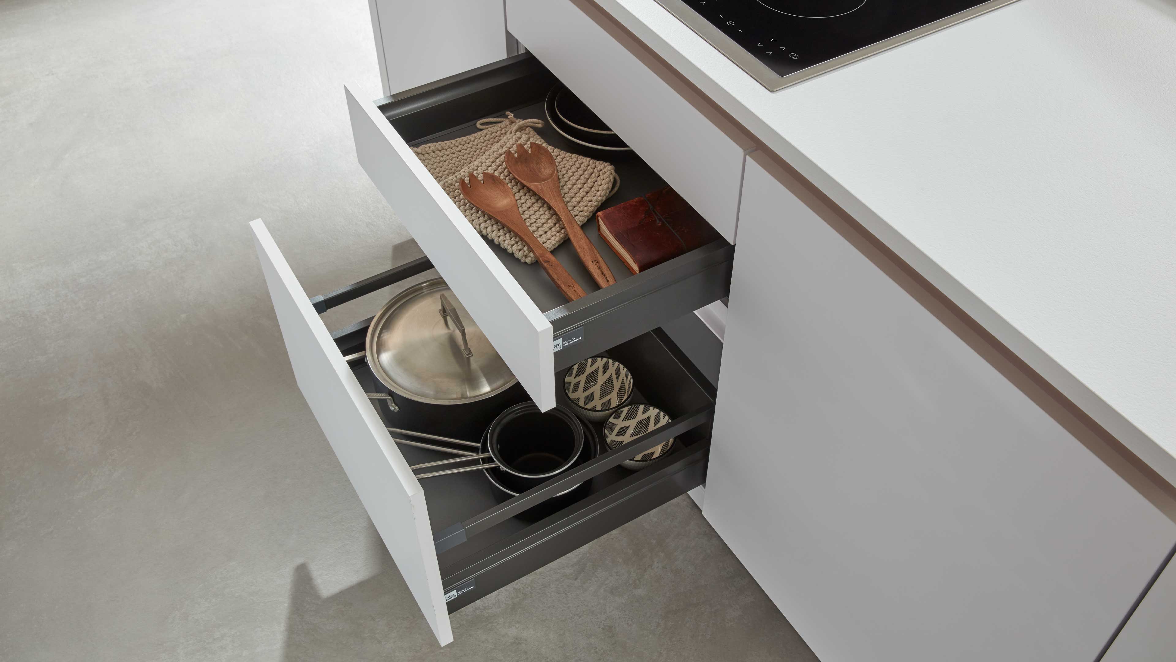 Weiße Küchenschubladen mit matten, grauen Innenkästen, obere Schublade mit Holzsalatlöffeln und Topflappen, untere mit Töpfen und Deckel.