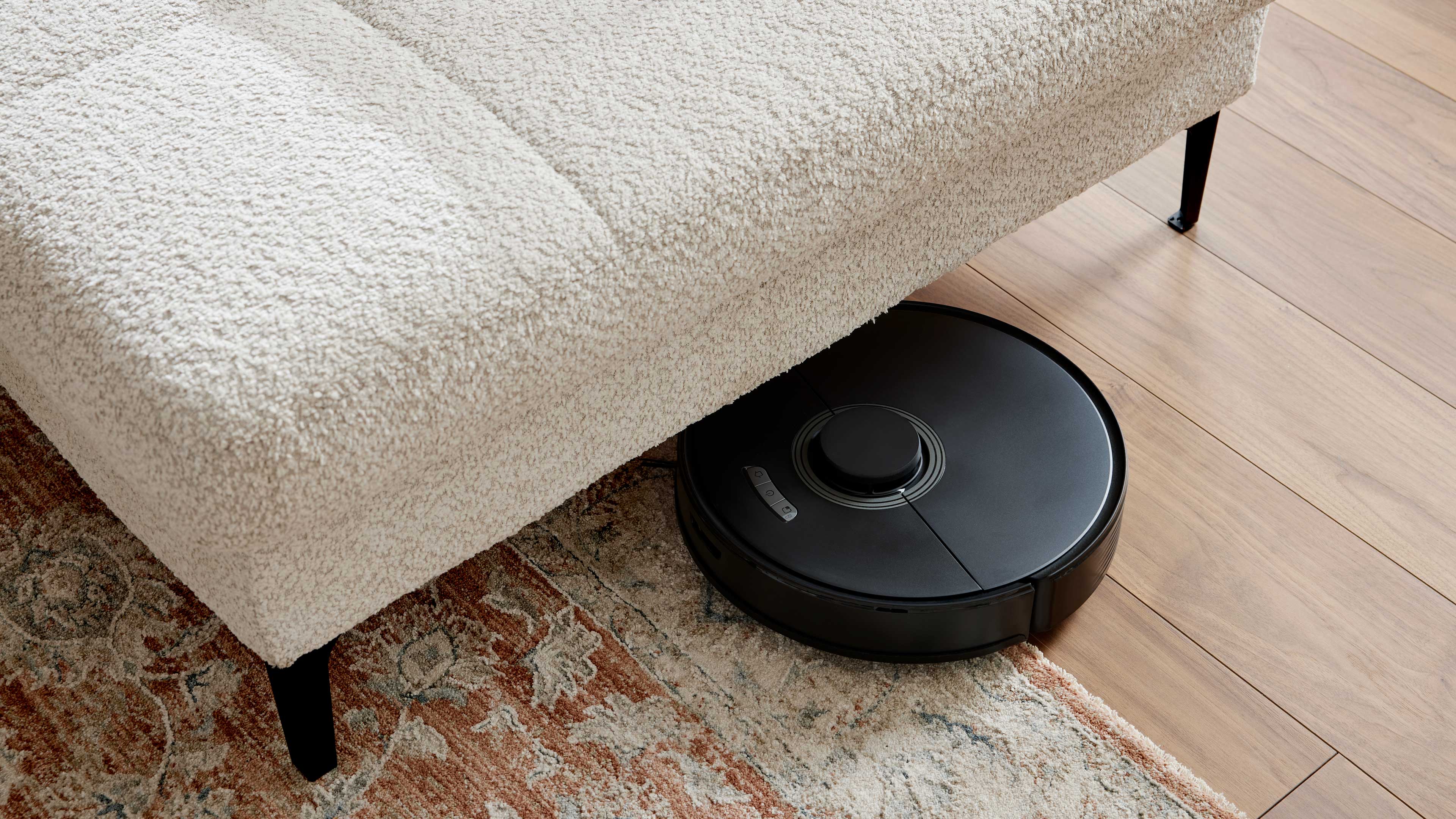 Beiger Bouclé-Sofaabschnitt mit schwarzen Metallbeinen, auf Holzboden, daneben ein schwarzer Saugroboter, Teil eines gemusterten Teppichs.