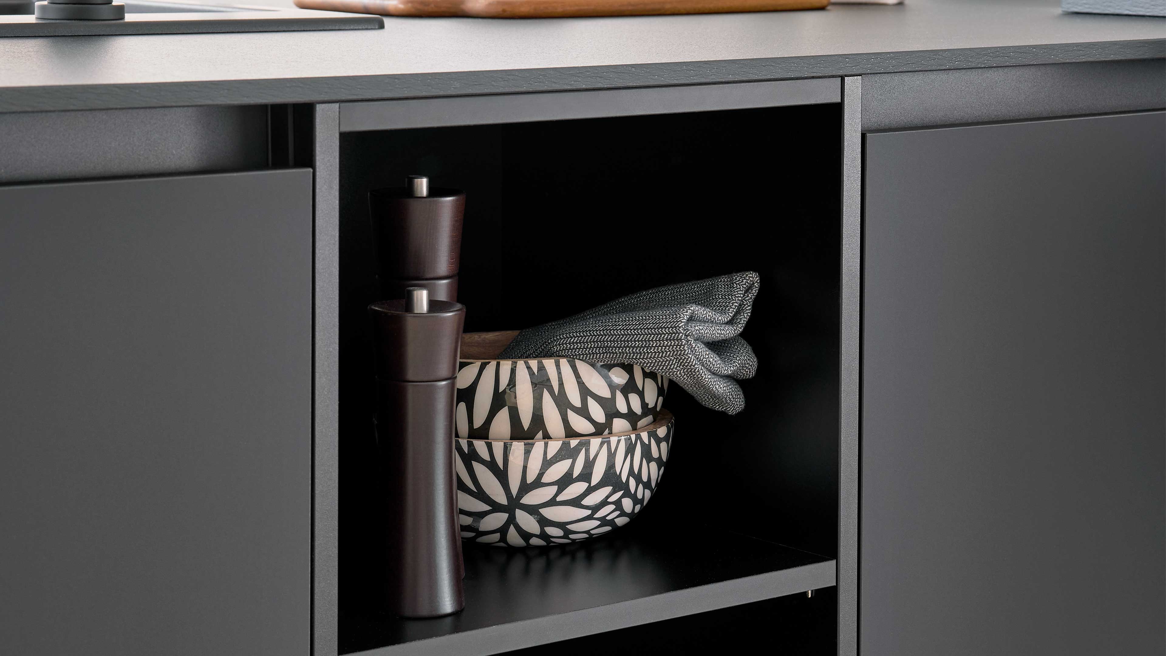 Offenes Fach eines dunkelgrauen Sideboards mit zwei schwarz-weiß gemusterten Keramikschalen, grauem Stofftuch und zwei dunklen Holzmühlen.
