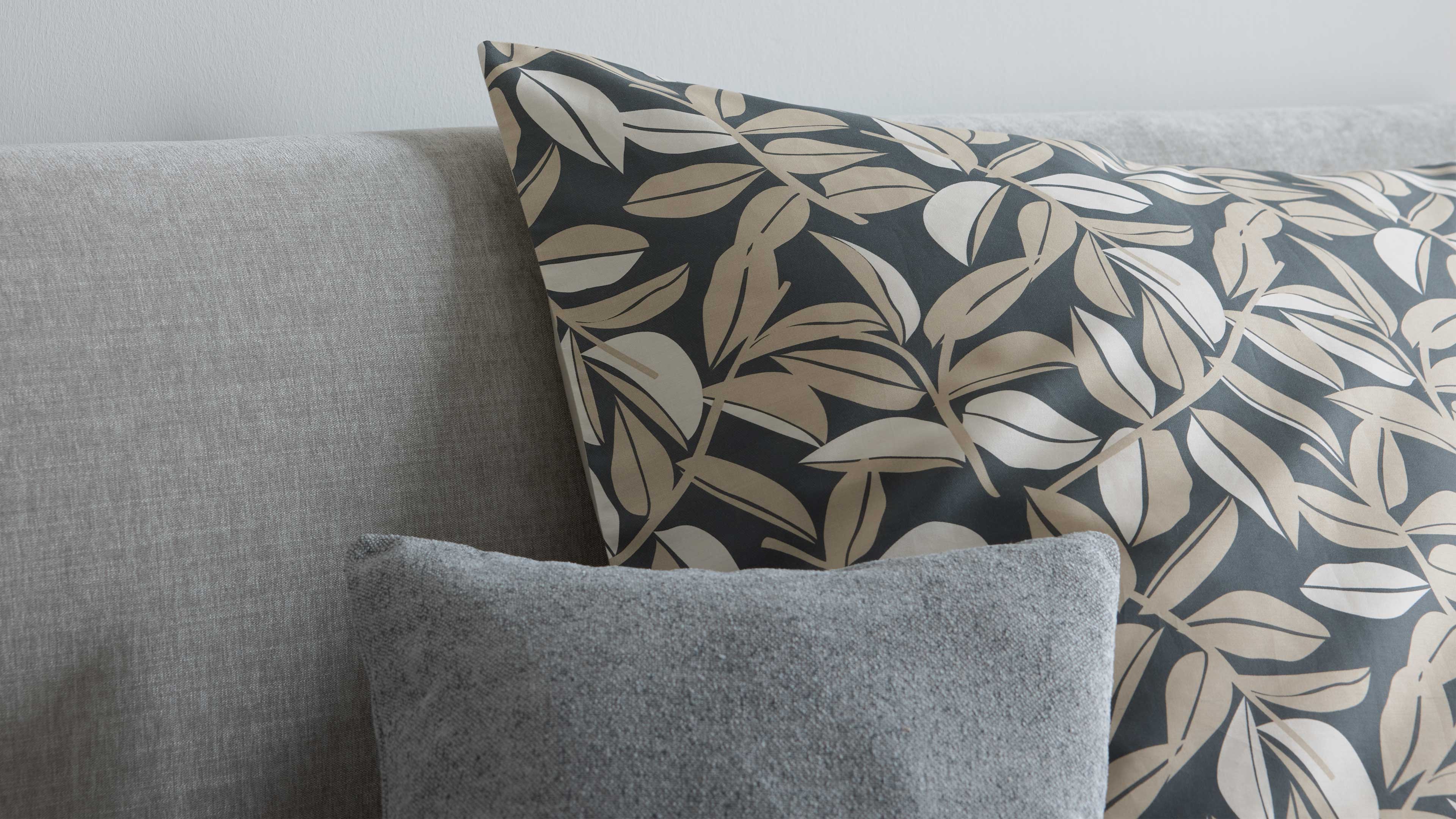 Großes Kissen mit dunklem Hintergrund und beigem Blattmuster lehnt an grauem Sofa, davor ein kleines hellgraues Strukturkissen.