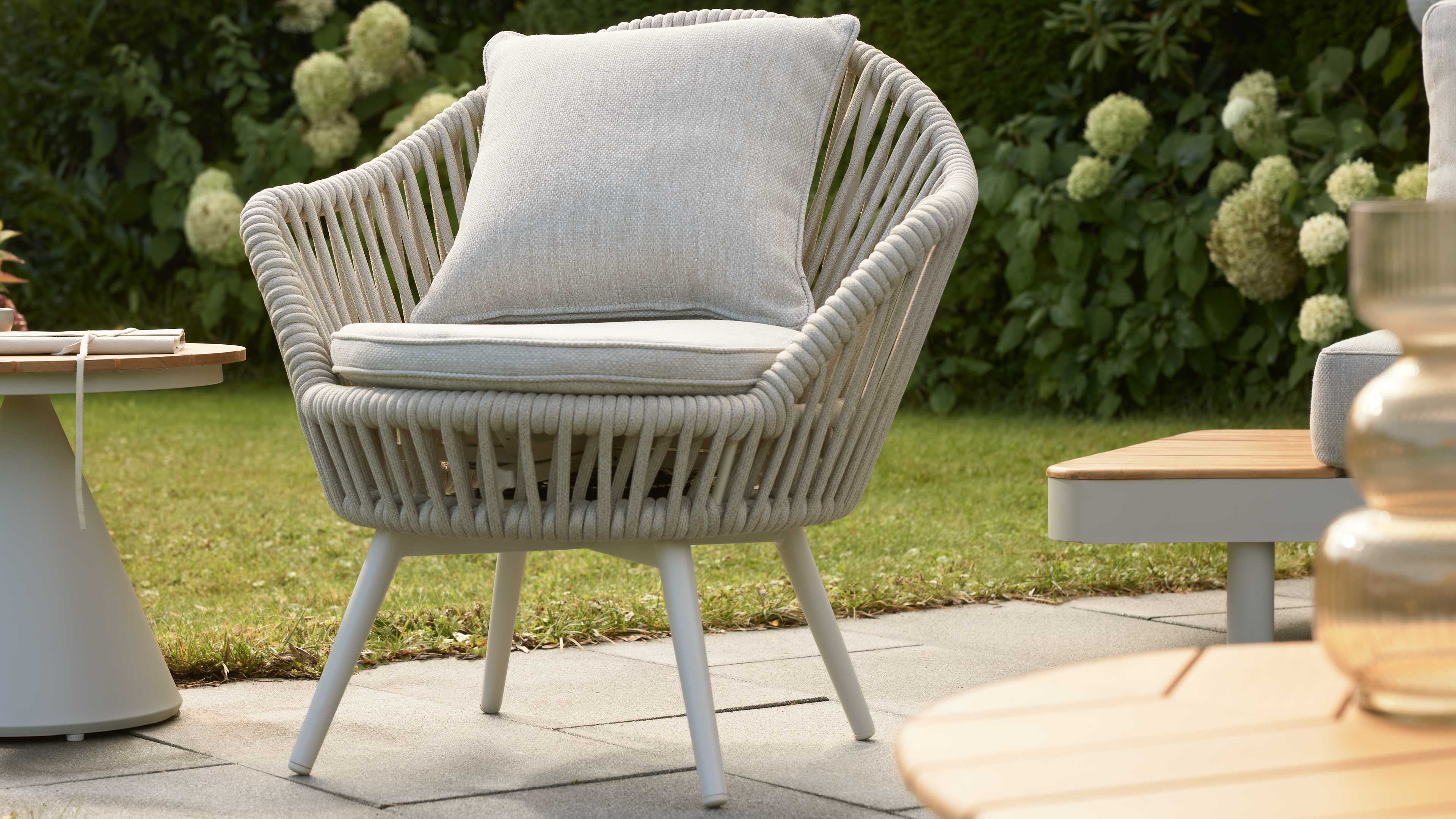 Rundgeformter Outdoor-Sessel mit beige-grauem Sitzkissen und geflochtenem Seilgestell, abgestellte Beistelltische, Rasen und Hortensien.
