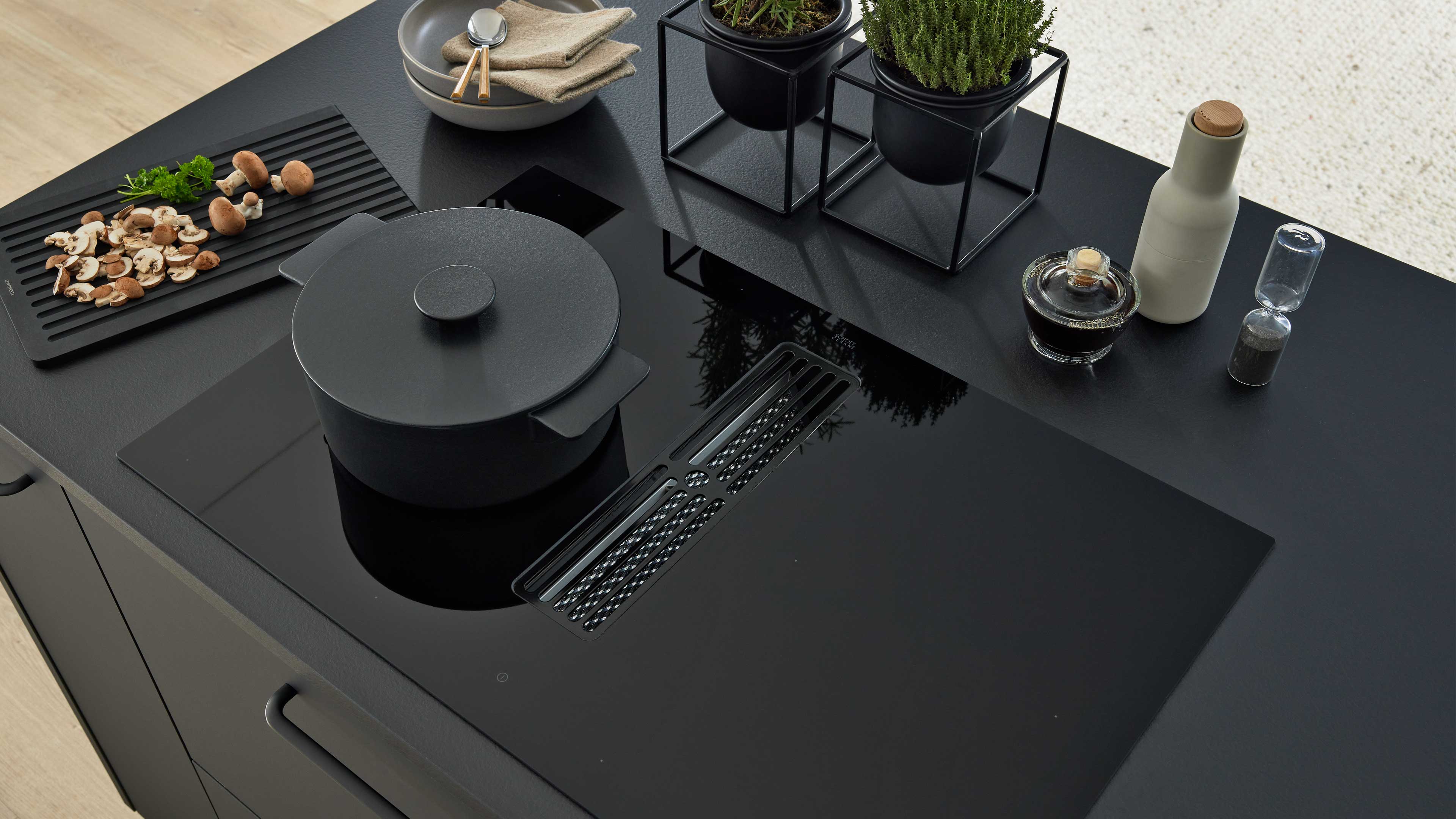 Schwarzes Kochfeld mit Lüftungsgitter und schwarzem Topf, links Schneidebrett mit geschnittenen Champignons, rechts zwei Kräutertöpfe.