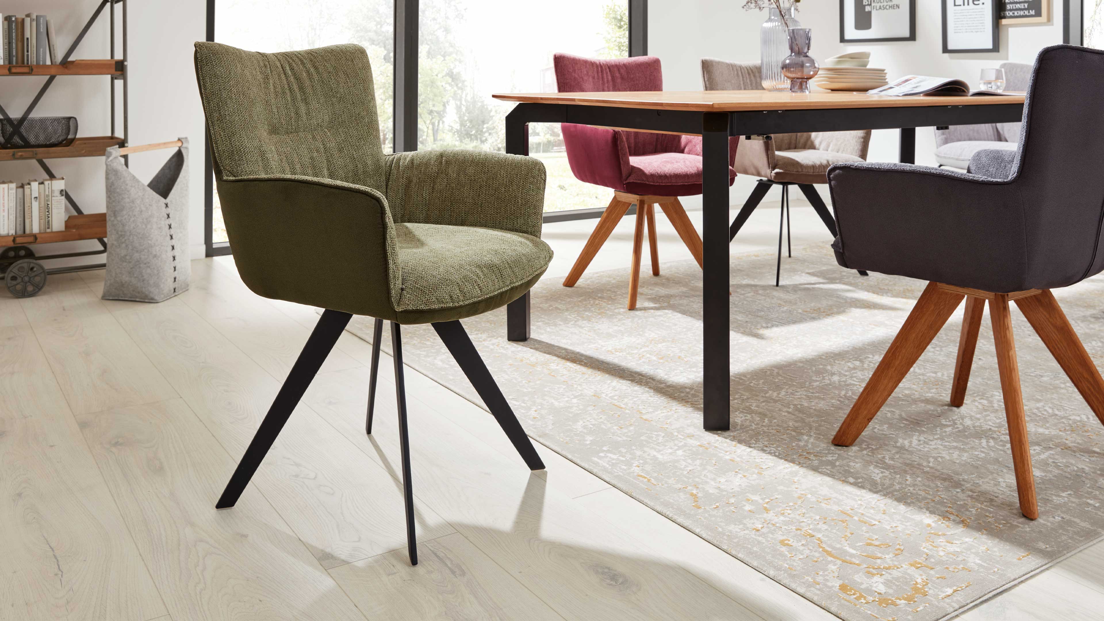 Grüner gepolsterter Armlehnstuhl mit schwarzem, vierbeinigem Metallgestell im Vordergrund, dahinter Esstisch, bunte Stühle und Teppich.