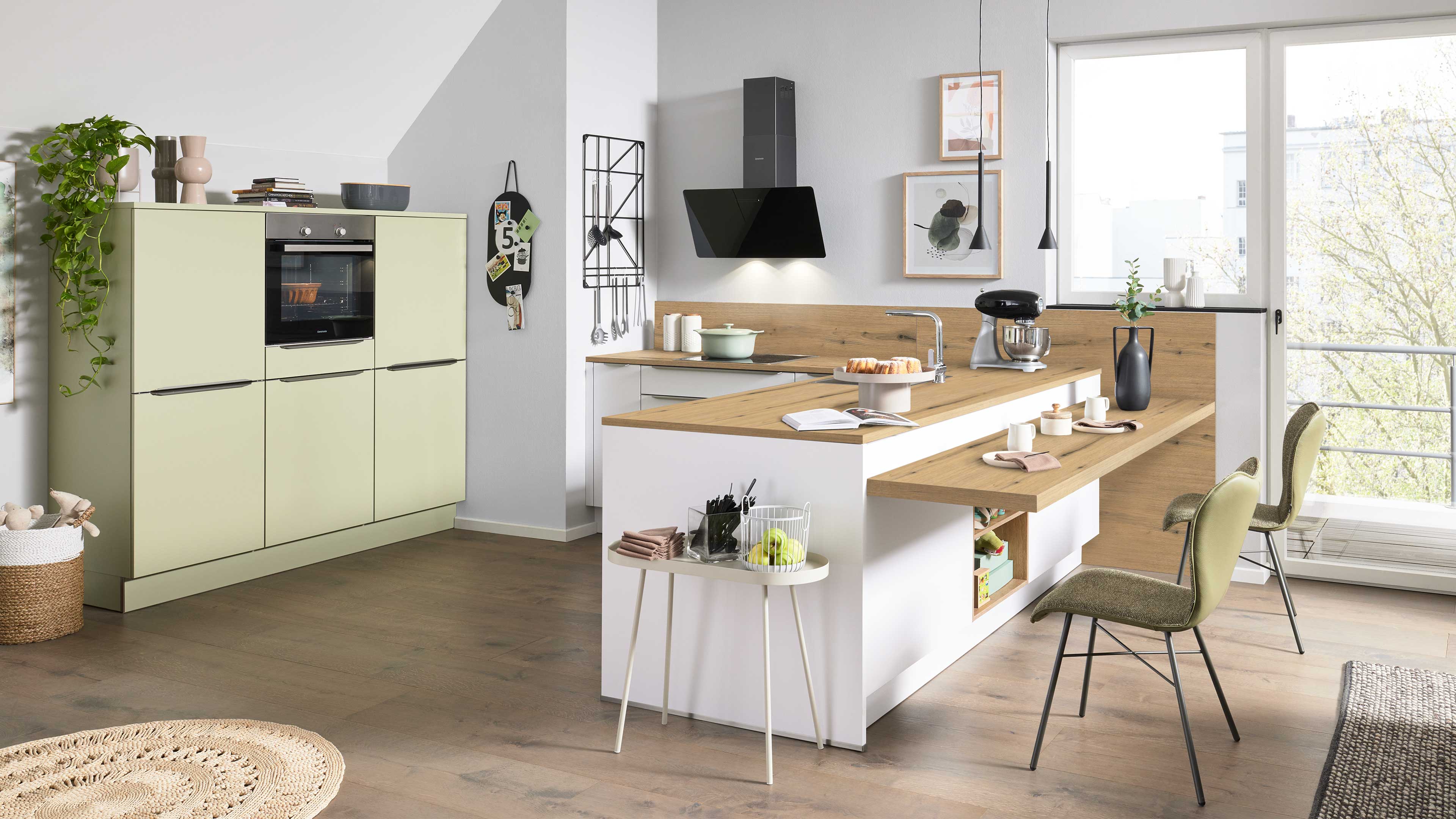 Kücheninsel mit Holzarbeitsplatte, Spülbecken, Tresen und zwei grünen Stühlen; links pastellgrüne Schränke mit Backofen.