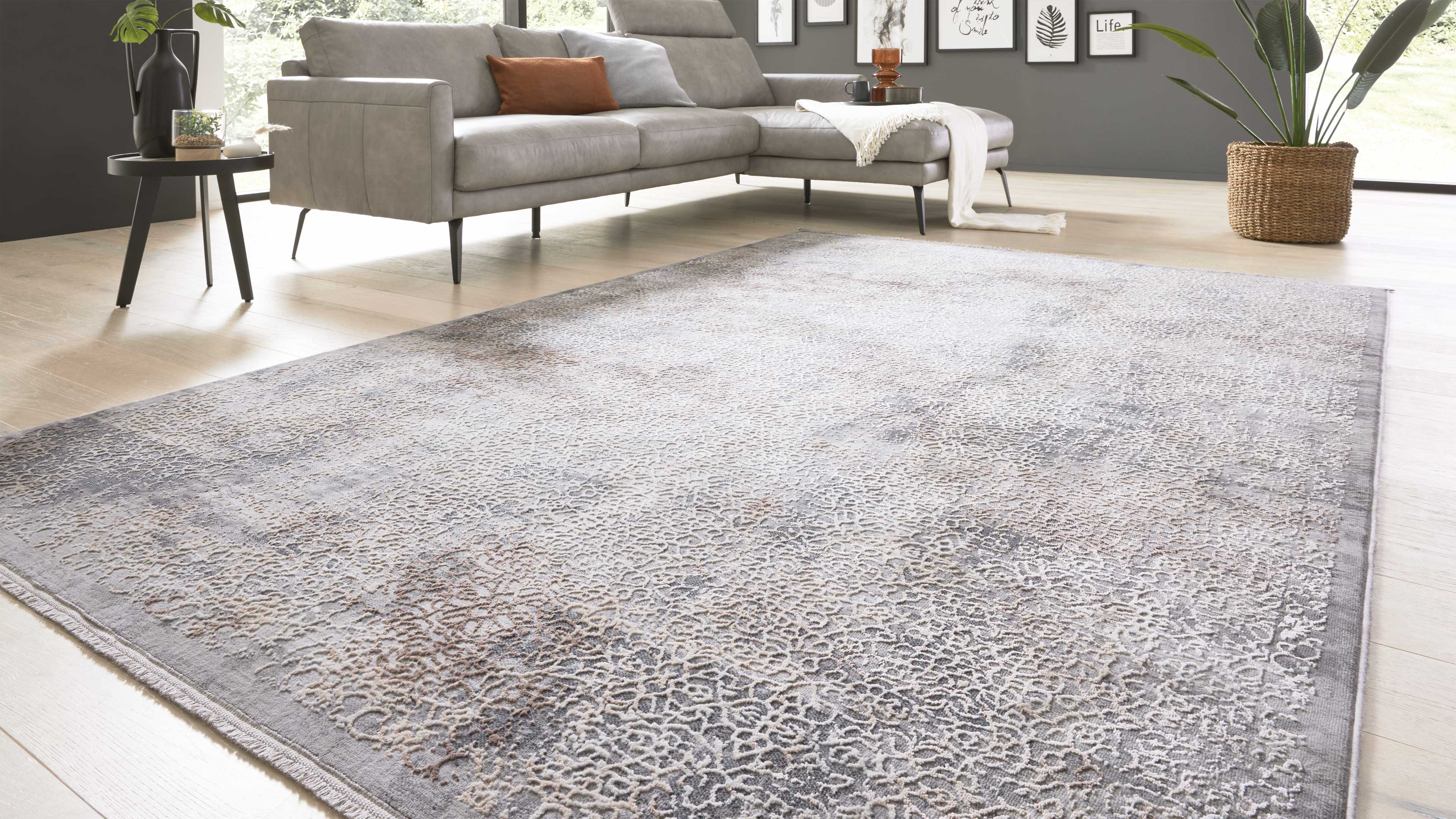 Großflächiger hellgrauer Teppich mit feinem, leicht ornamentiertem Muster, davor eine graue Eckcouch, Beistelltisch und Pflanzen.