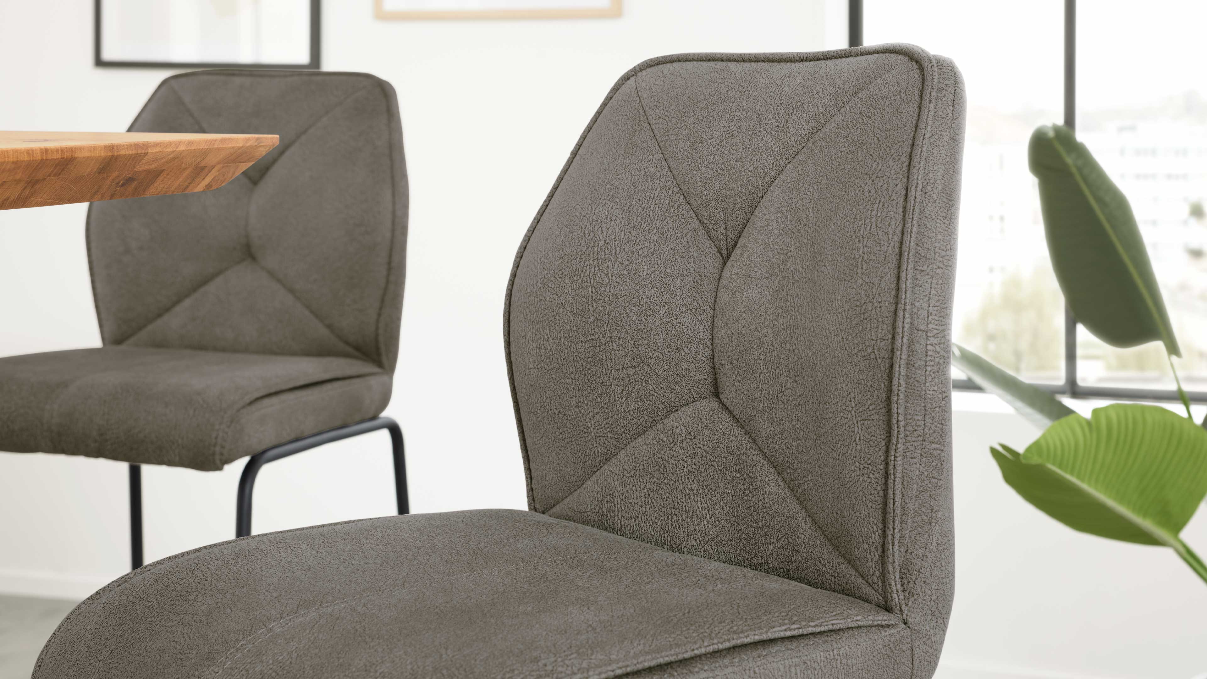 Grau bezogener Stuhl mit strukturierter Oberfläche im Vordergrund, dahinter ein zweiter Stuhl, Tischkante links und Grünpflanze rechts.