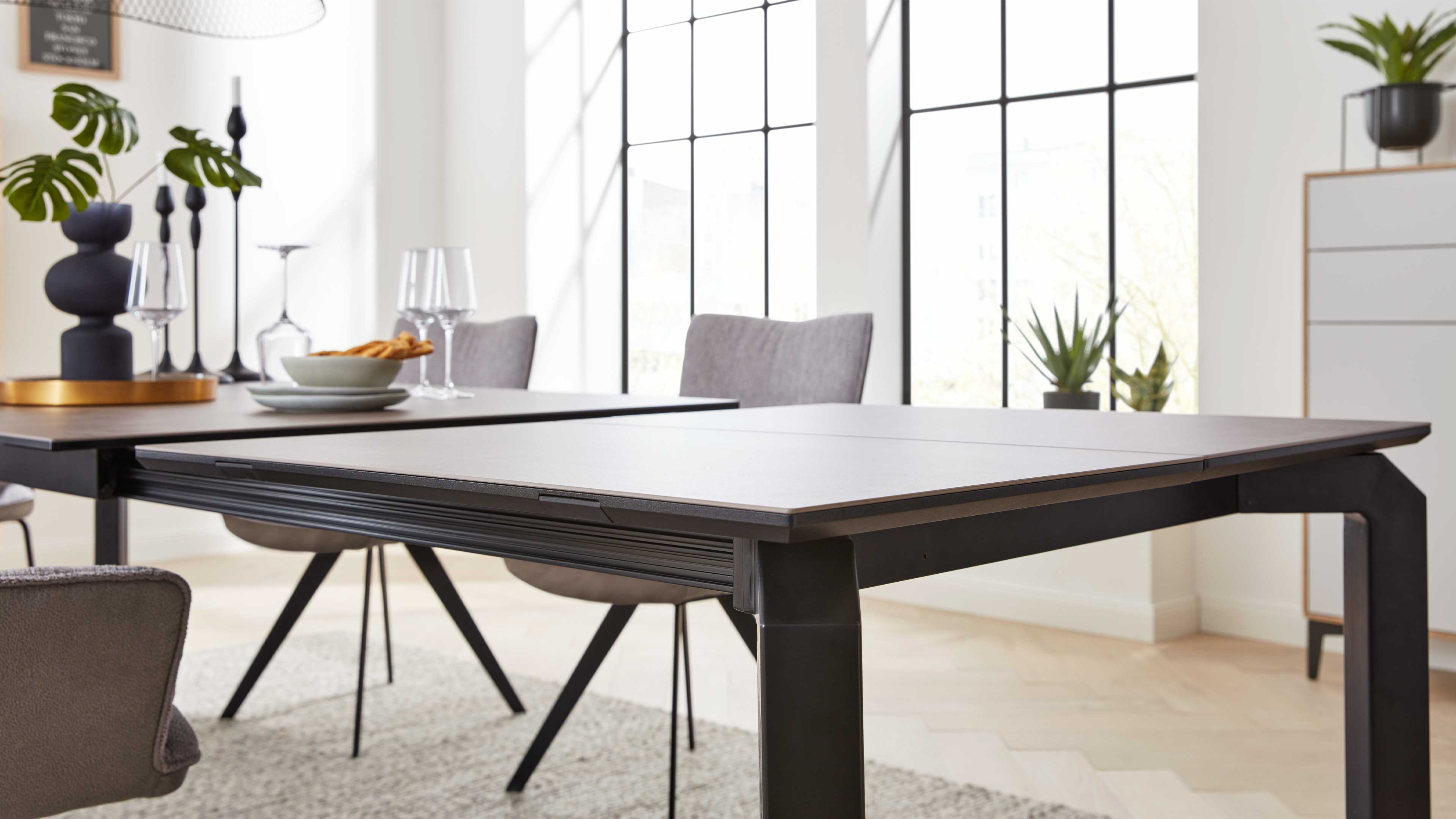 Rechteckiger Esstisch mit dunklem Metallgestell und hellem, zweiteiligem Tischblatt, umgeben von grauen Stühlen und großen Fenstern.