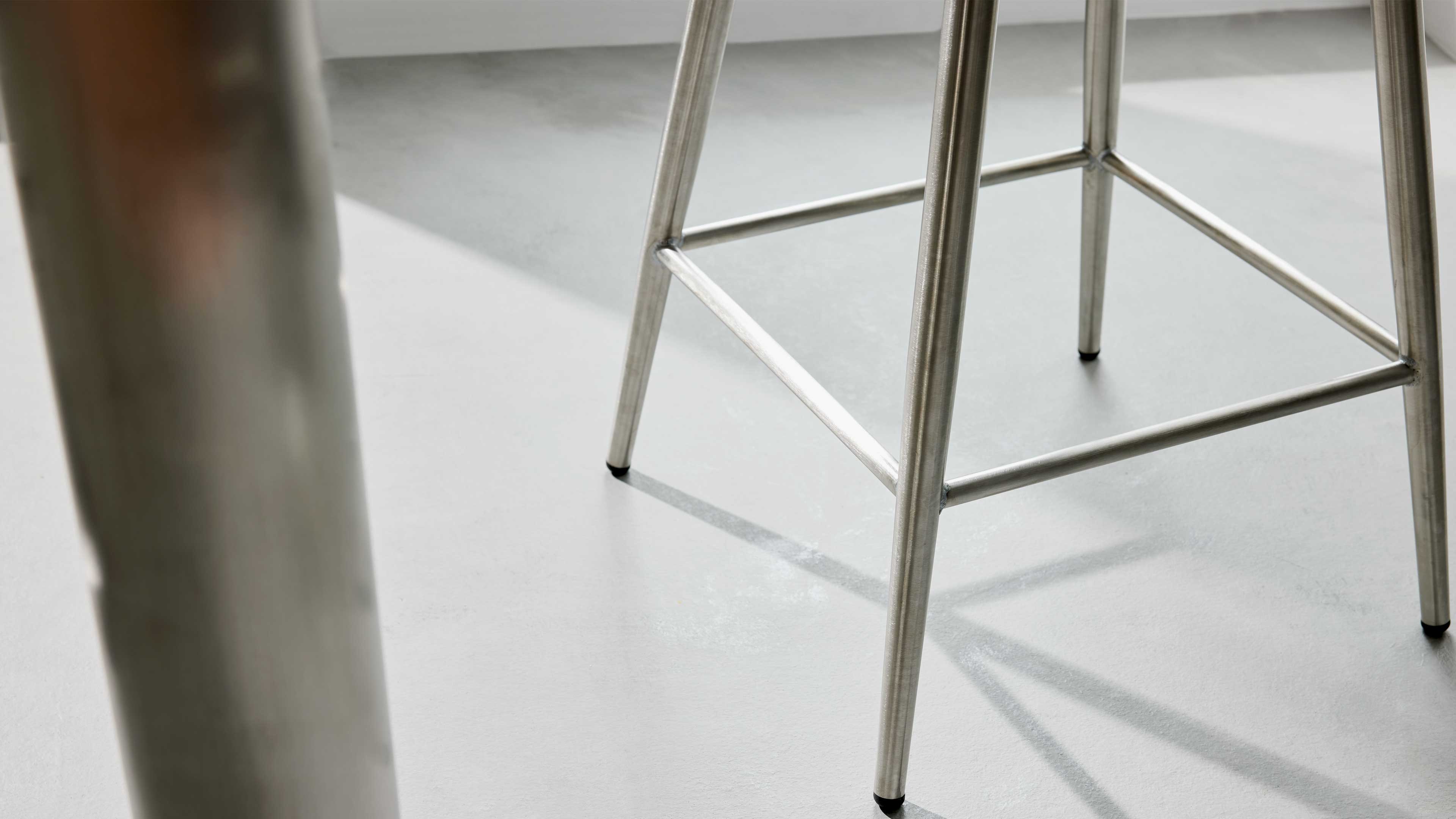 Mehrere runde Metallbeine eines Hockers mit Querstreben und kleinen Gummikappen stehen auf hellem Boden, Schatten sichtbar.