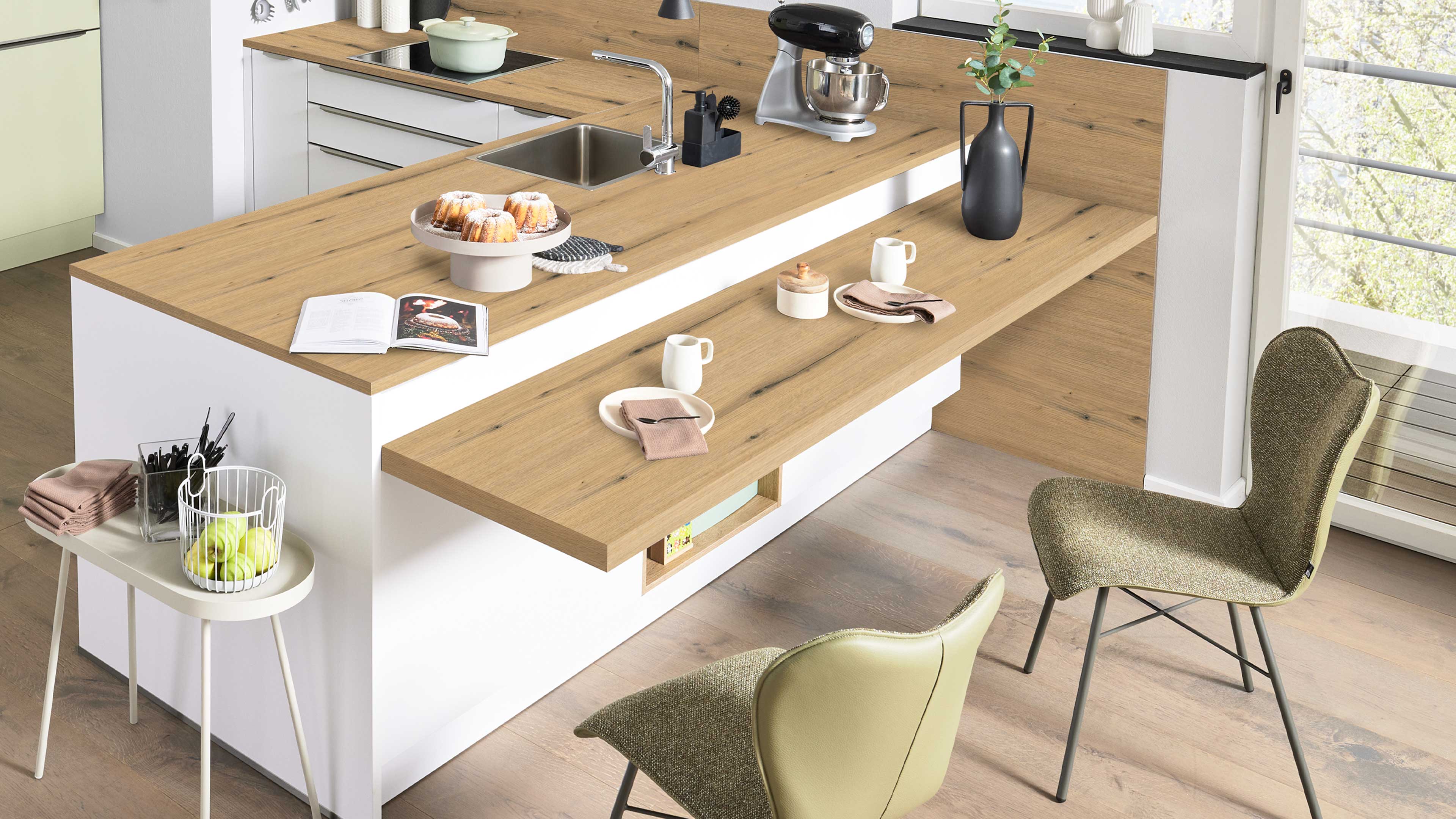Kücheninsel mit heller Holzarbeitsplatte und aufgesetzter Theke, integriertem Spülbecken, Standmixer, Tellern, Vase und zwei grünen Stühlen.