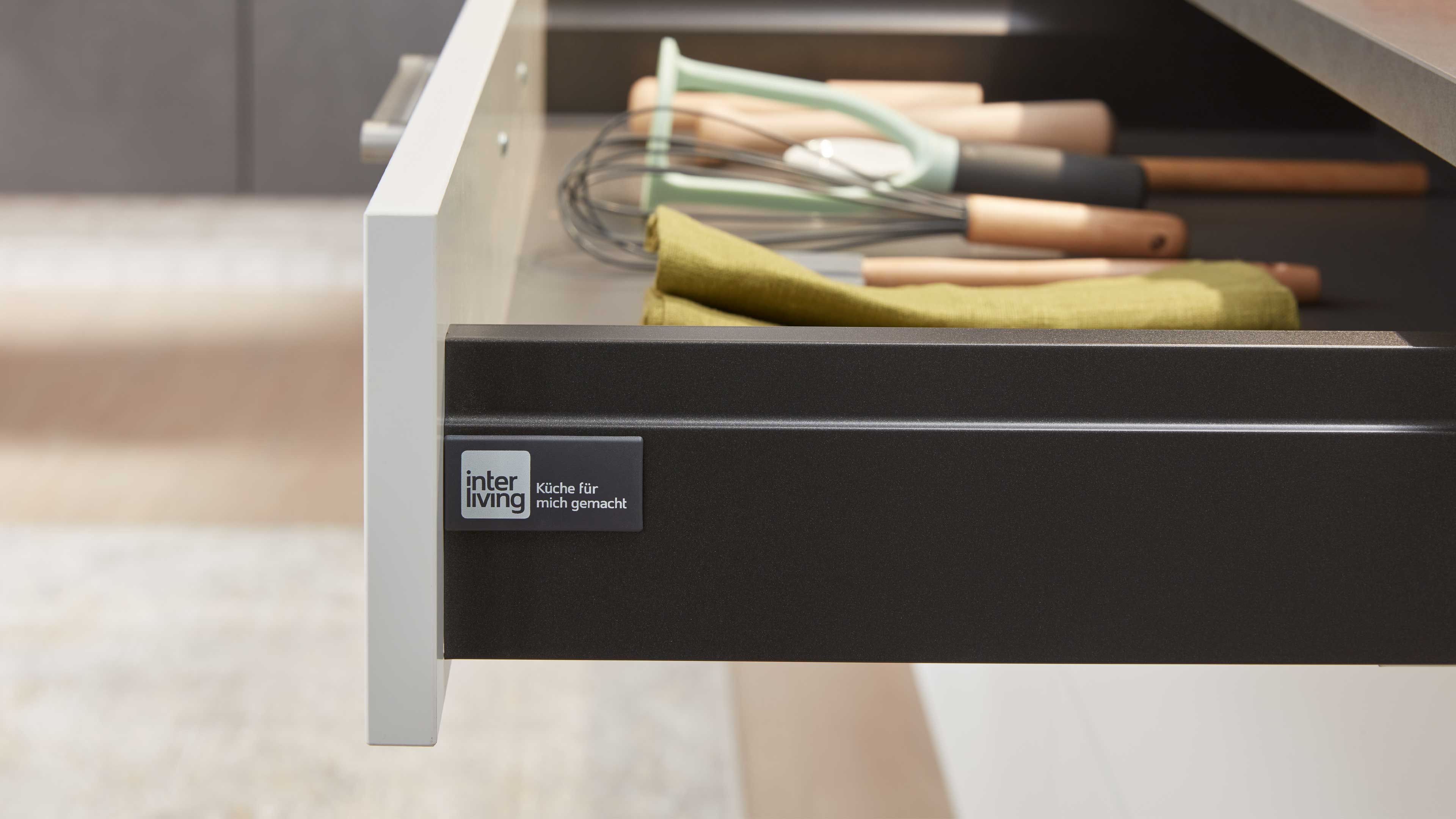 Geöffnete Küchenschublade mit weißer Front und dunkelgrauem Innenraum, Holzgriff-Utensilien, grünes Tuch und Markenplakette.