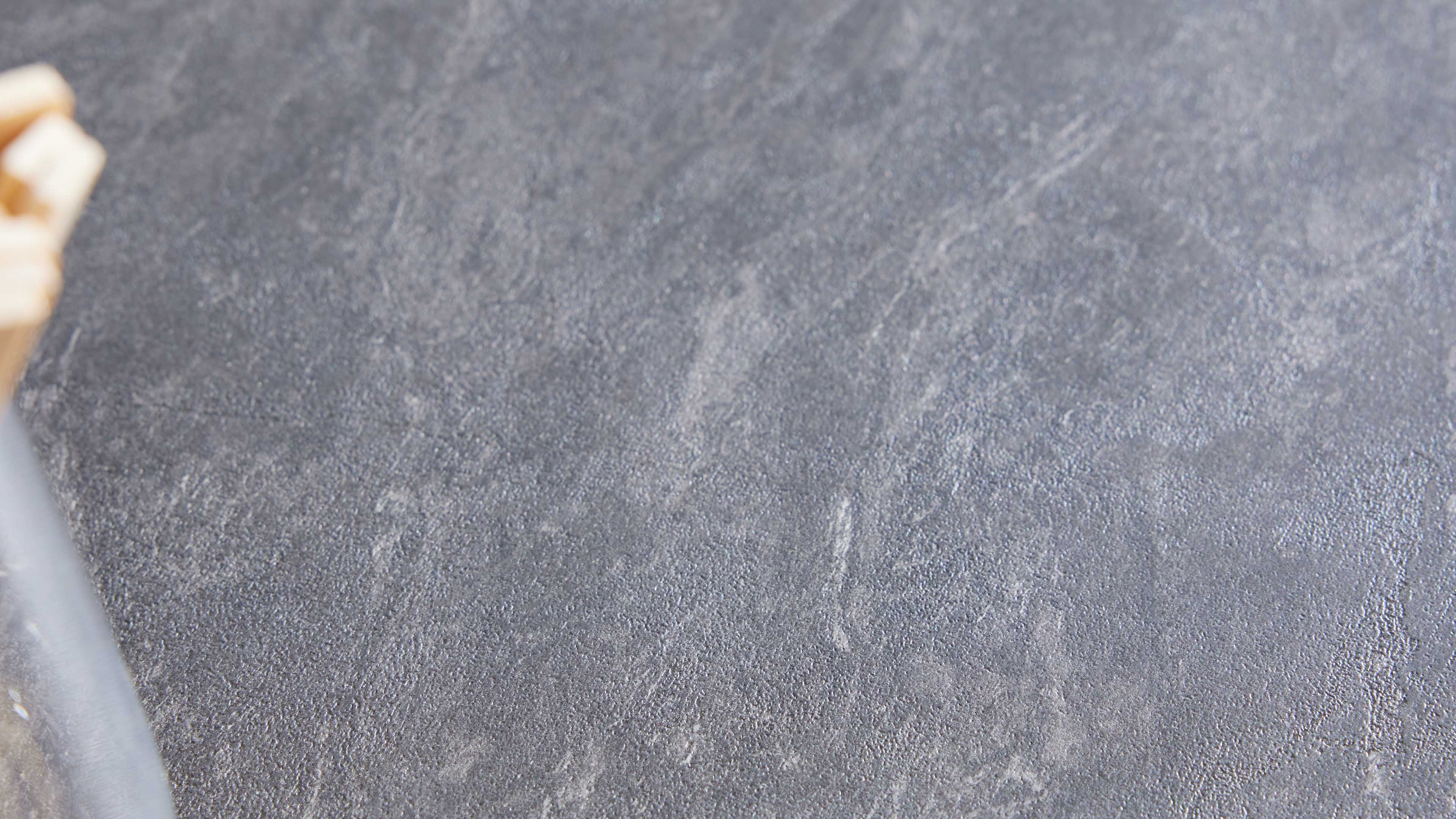 Dunkelgraue, leicht strukturierte Tischplatte mit feinen weißen Aderungen; links unscharf Holzdetail und dunkler Stab.