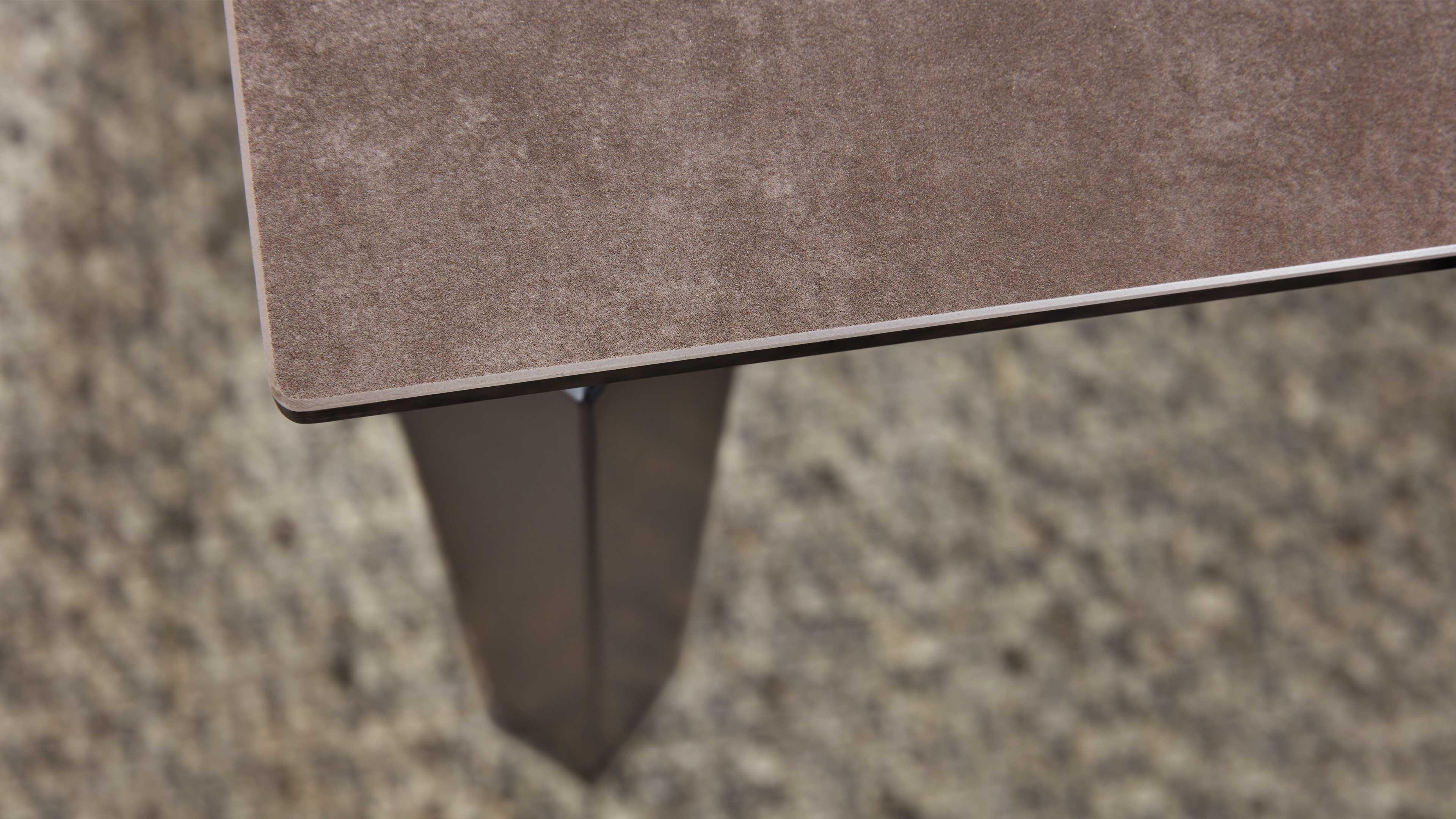 Eckdetail einer rechteckigen Tischplatte mit brauner samtiger Oberfläche und sichtbarem trapezförmigem Metallbein über beigem Teppich.