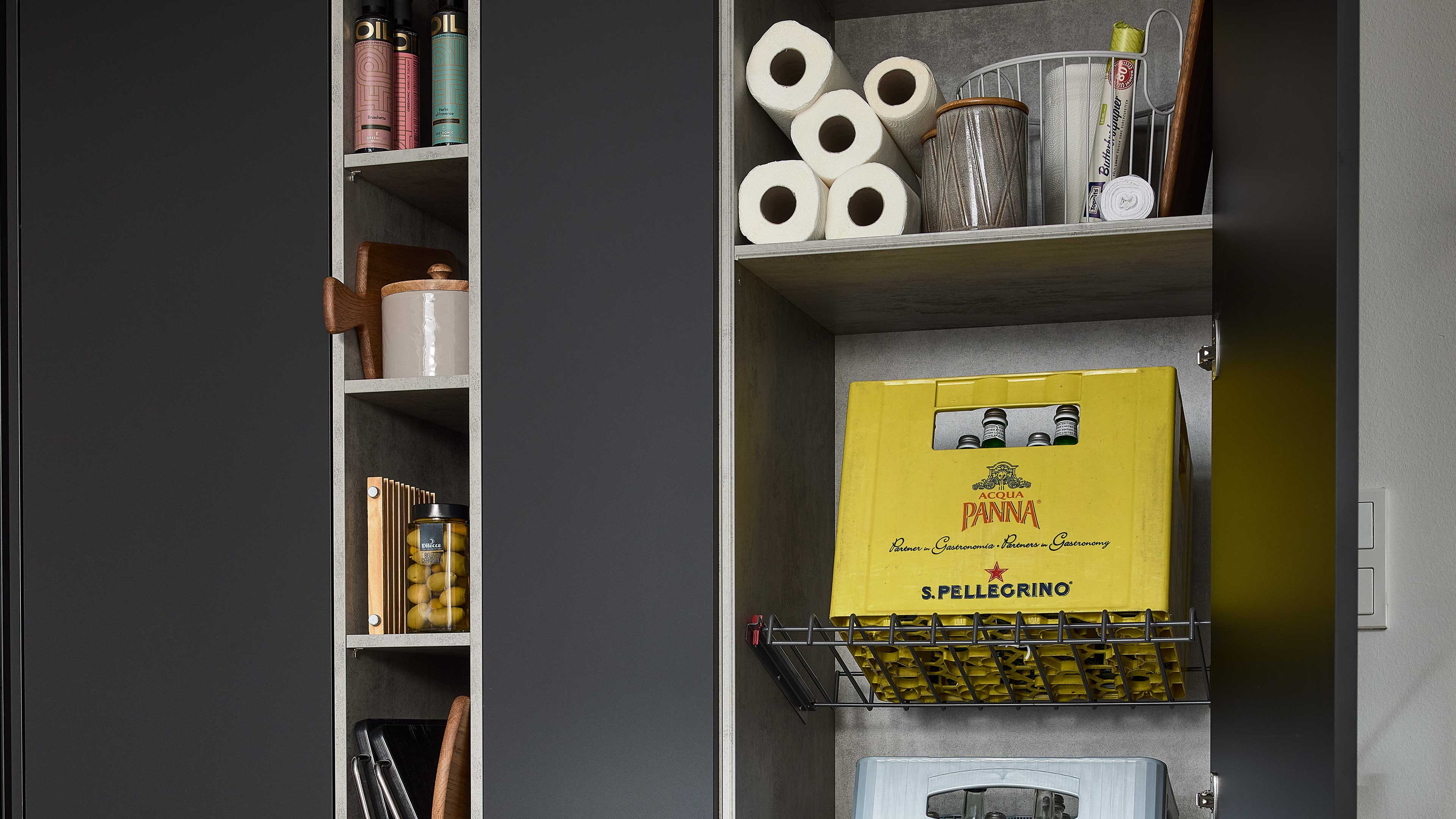Offener grauer Küchenschrank mit gelber Getränkekiste auf einem ausziehbaren Drahtfach, darüber mehrere Toilettenpapierrollen und Dosen.