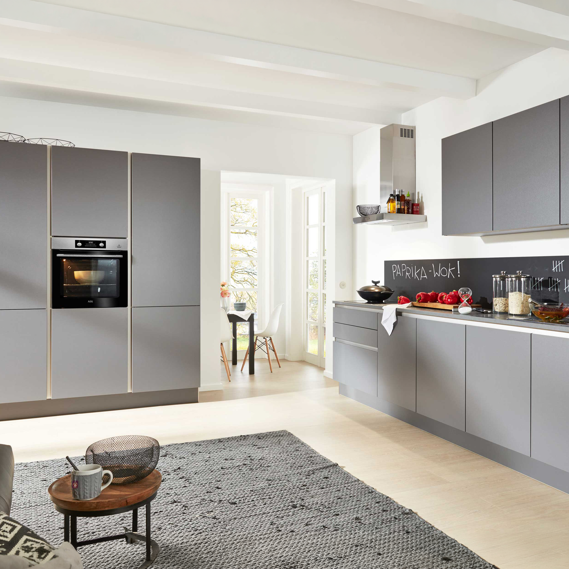 Offene Küche mit mattgrauen, griffarmen Fronten, Hochschrank mit Einbauofen, Hängeschränken und schwarzer Tafelfläche als Spritzschutz.