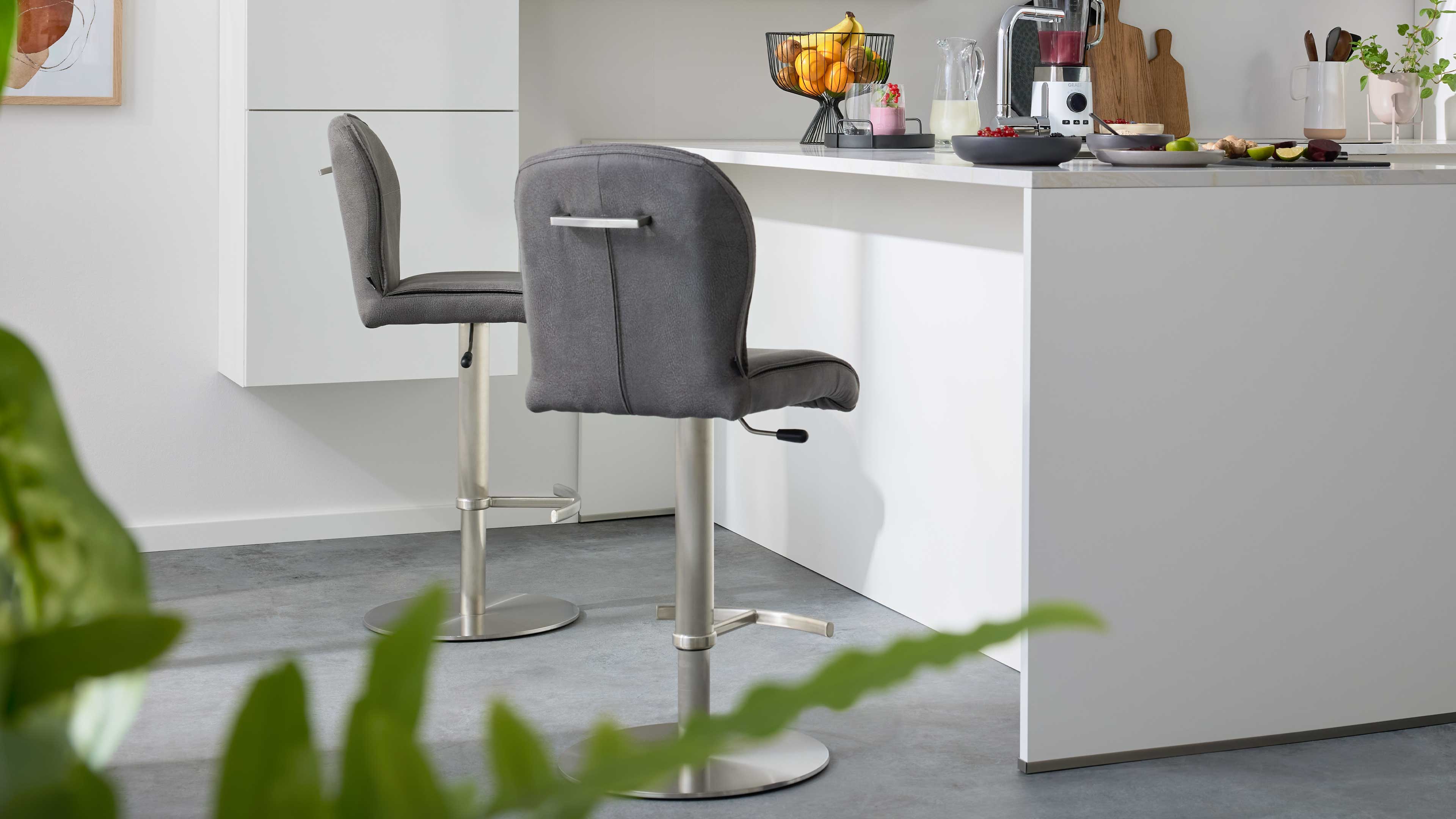 Zwei graue gepolsterte Barhocker mit Edelstahlfuß, Fußstützen, Höhenmechanik und Rückenhaltegriff vor weißer Kücheninsel mit Obstschale.