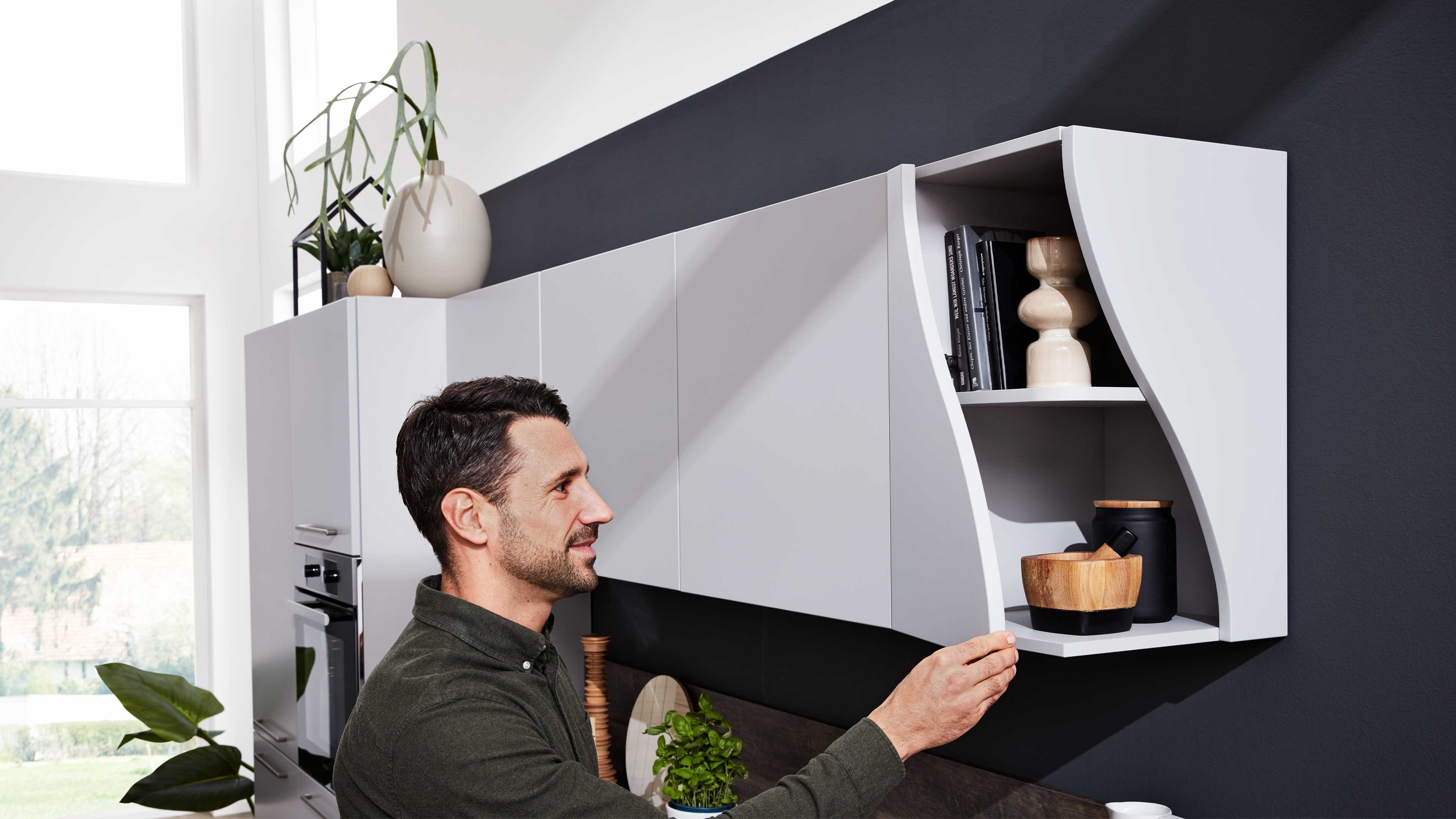 Ein Mann steht vor weißen Hängeschränken in einer Küche und öffnet ein offenes Regal mit Holzschalen, Büchern und Dekoration.