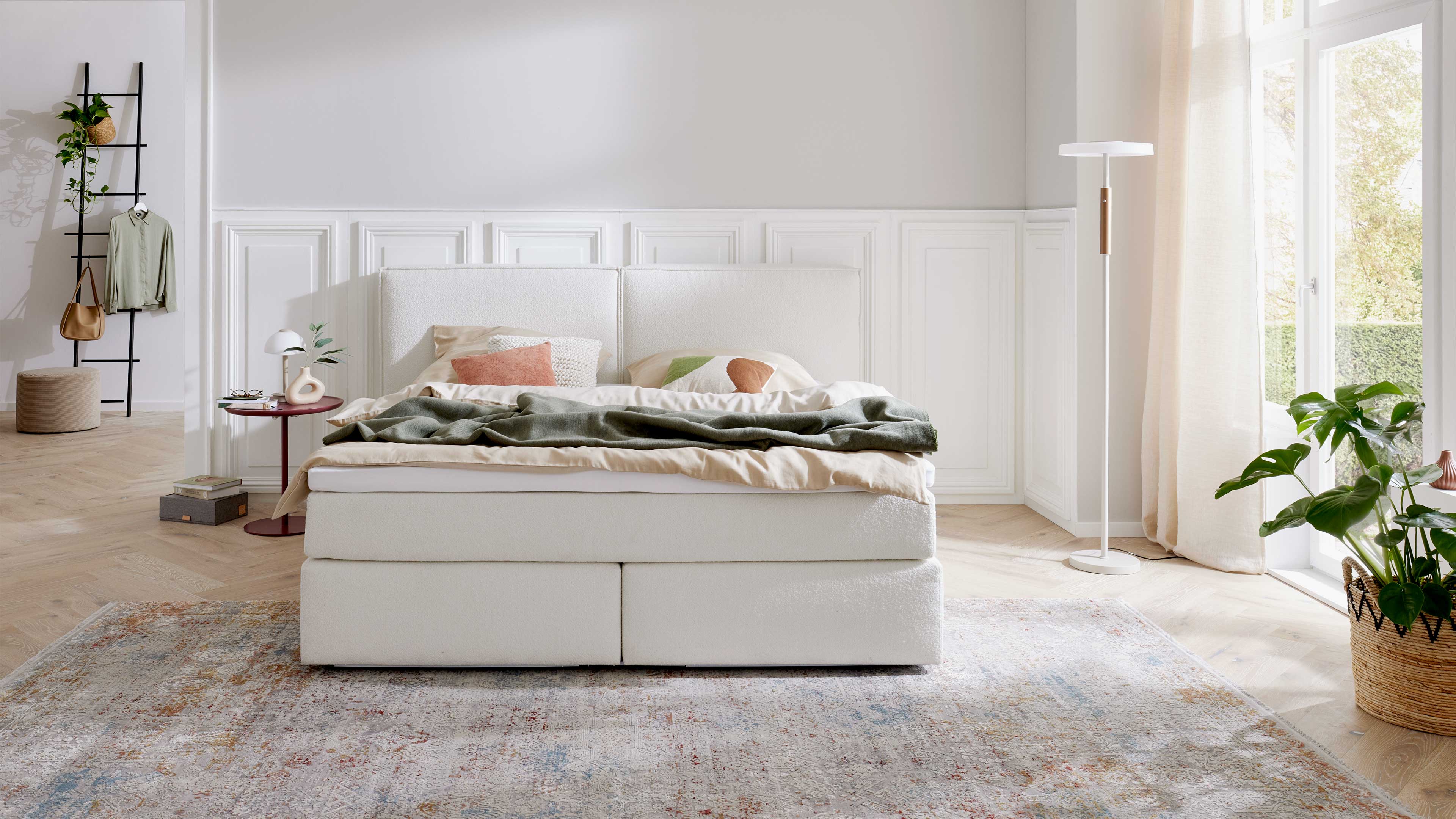 Weißes Polsterbett mit hoher Kopfteilfläche, beiger Bettwäsche, grüner Decke und dekorativen Kissen vor heller Wandvertäfelung.