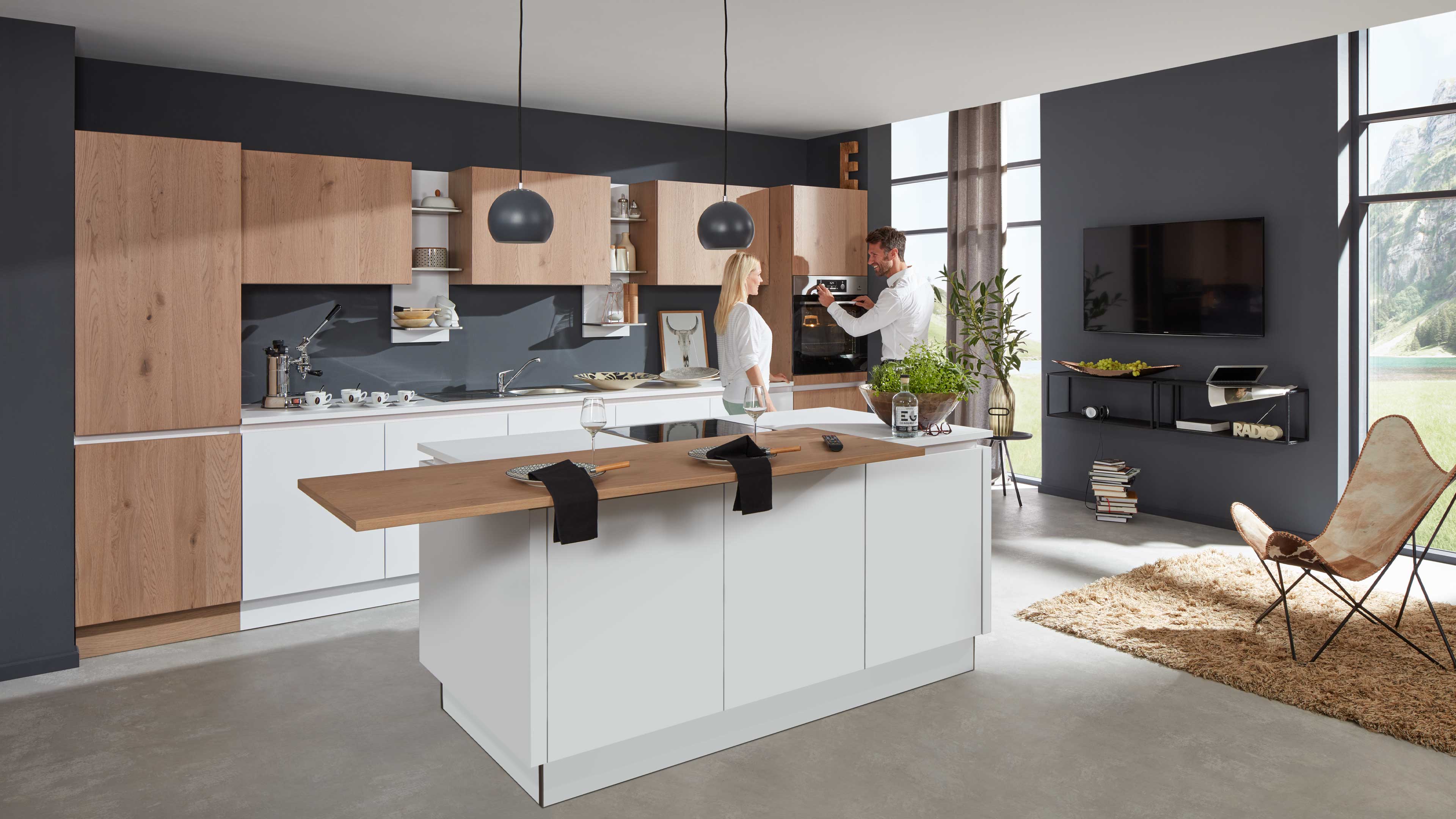 Küche mit weißer Insel und aufgesetzter Holzarbeitsplatte, dazu Wandküche mit Holzfronten und grauer Rückwand.