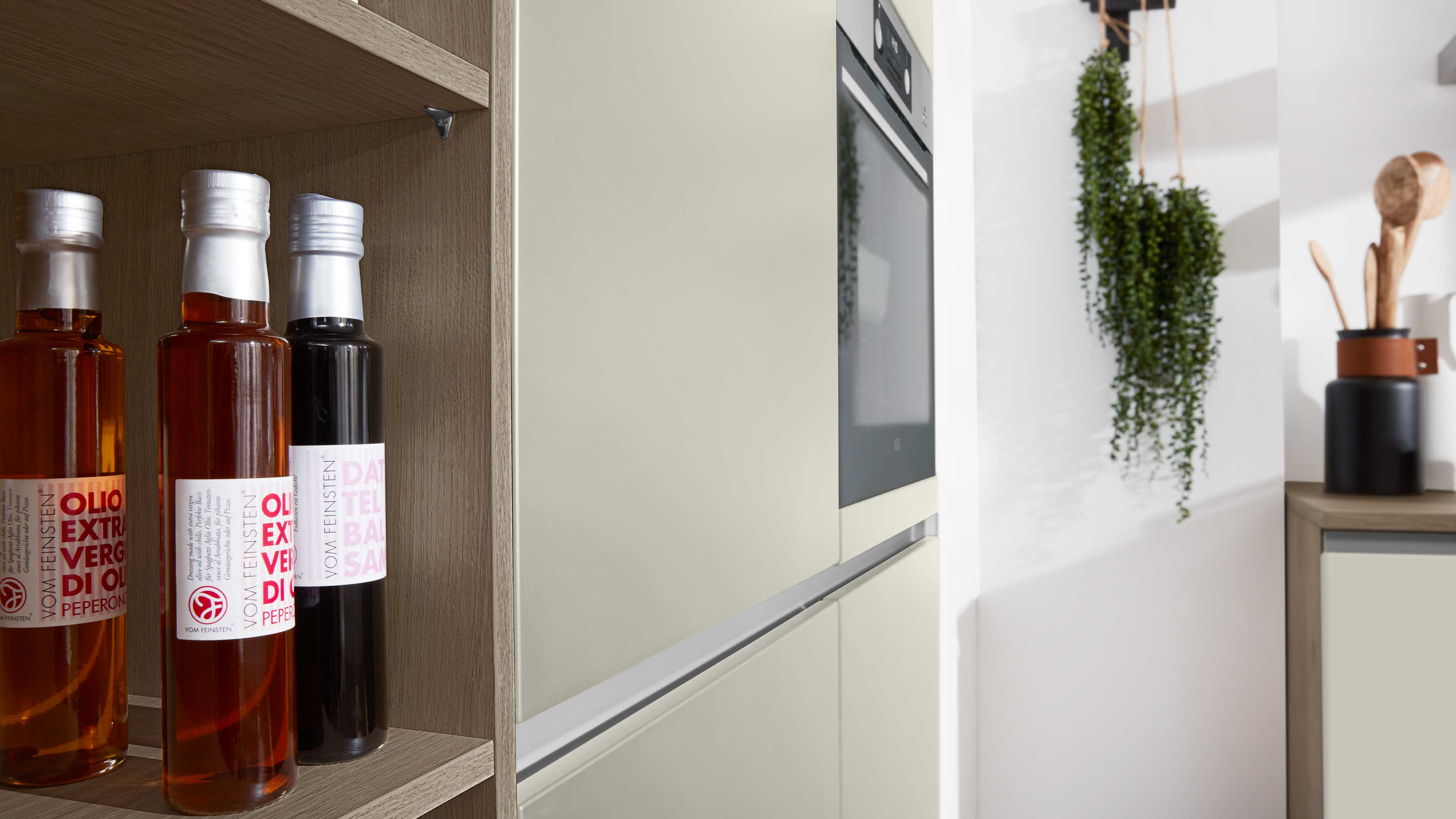 Offenes Holzregal mit drei Glasflaschen links, daneben beige Schränke mit Einbauofen, hängende Pflanze und Utensilien.