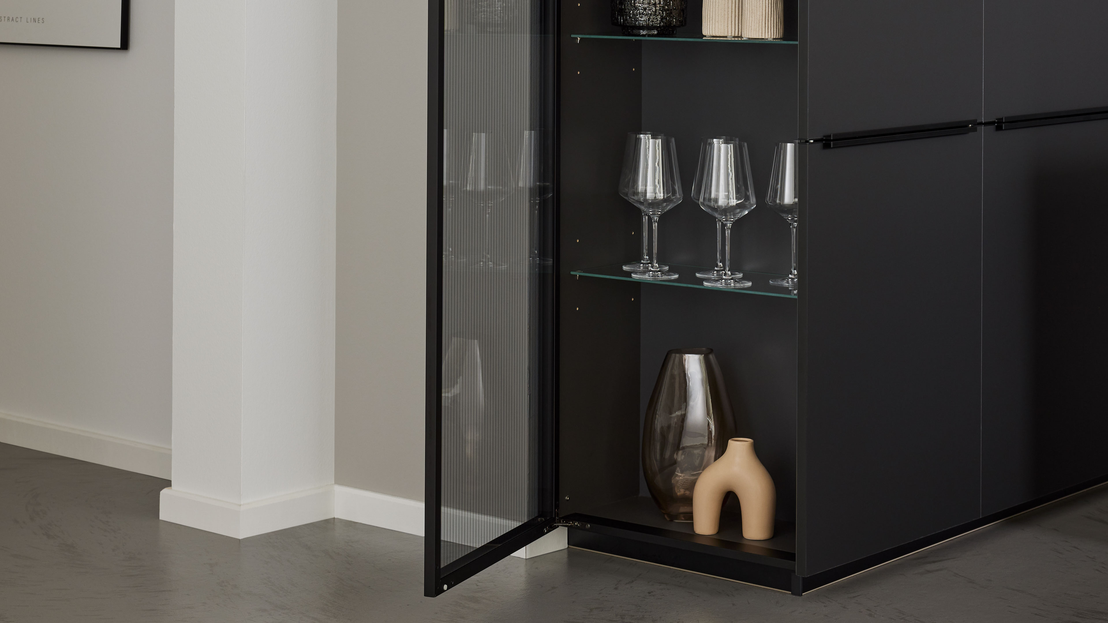 Schwarzer Schrank mit geöffneter, gerillter Glastür, drei Weingläsern auf einer Glasablage und zwei Vasen im unteren Fach.
