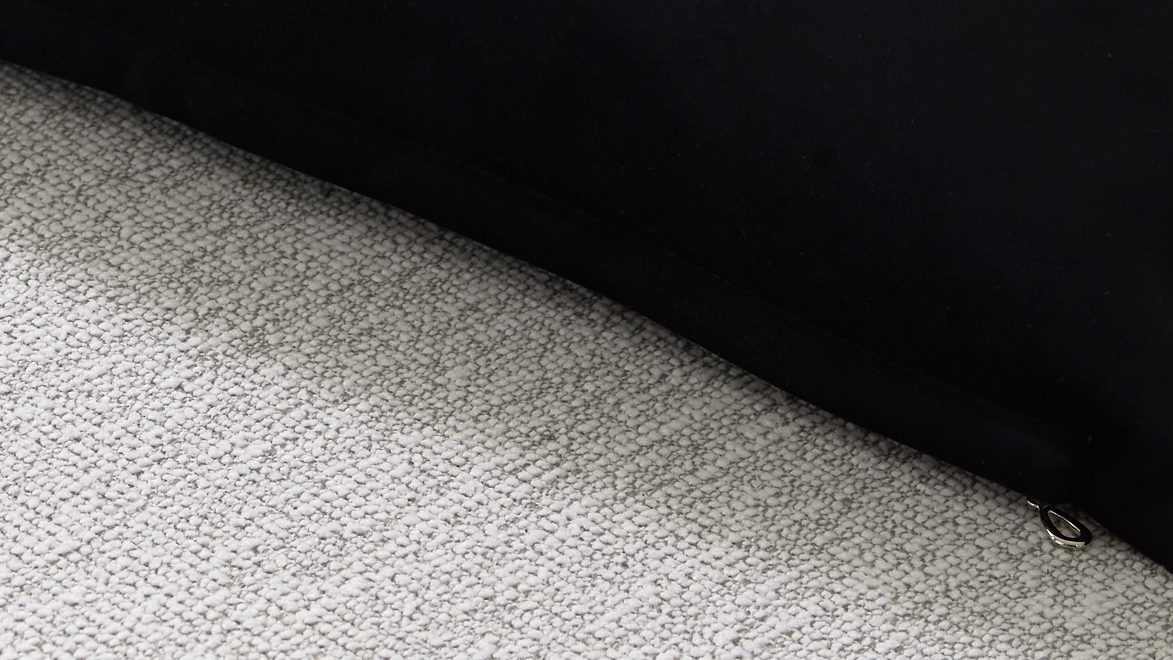 Schwarze Polsterkante auf hellgrauem, texturiertem Teppich mit sichtbarem kleinen metallischen Ring am unteren Bildrand.