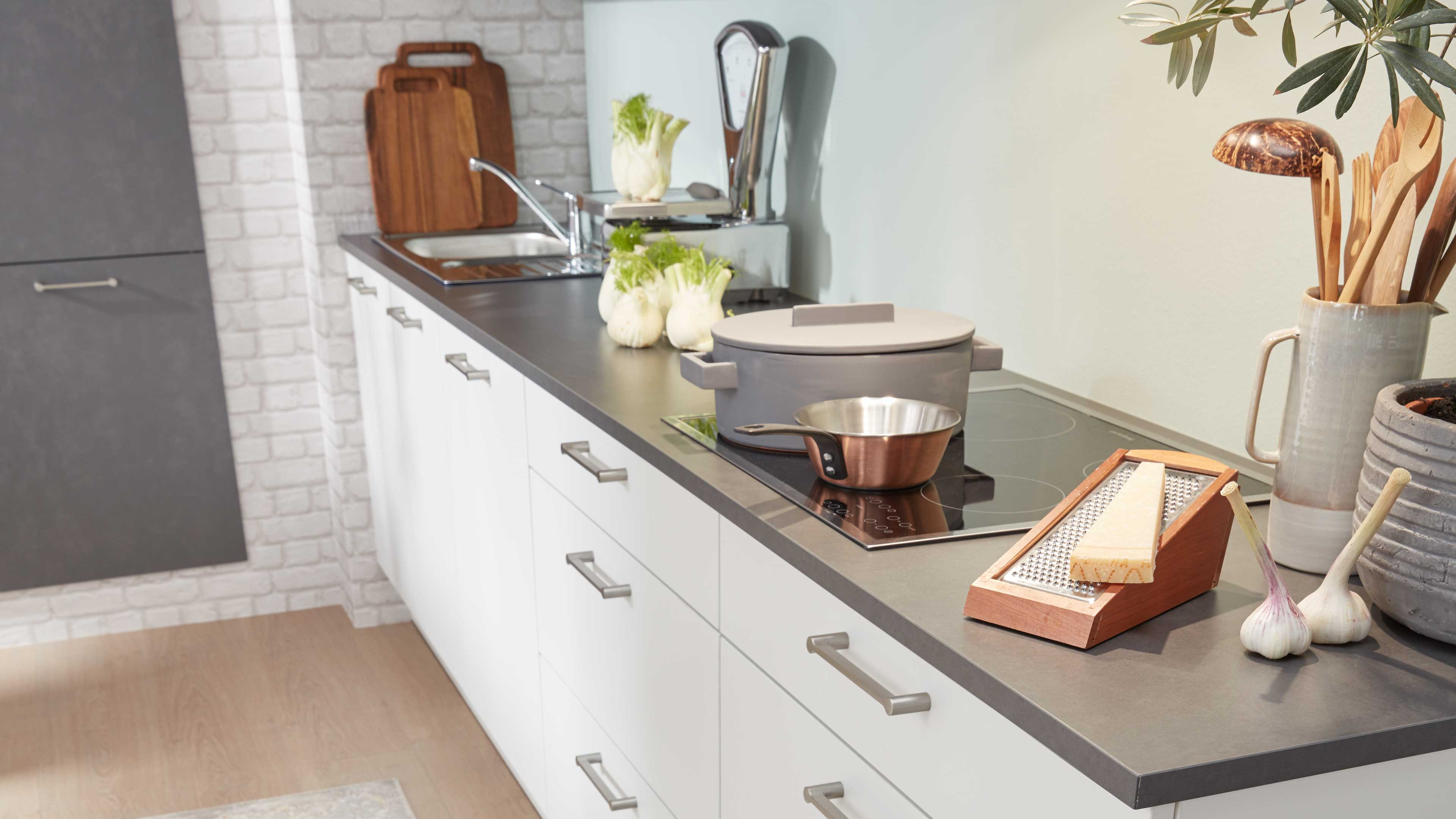Weißer Küchenunterschrank mit matter grauer Arbeitsplatte und Glaskeramik-Kochfeld, darauf grauer Topf, Kupferpfanne und Holz-Käsereibe.