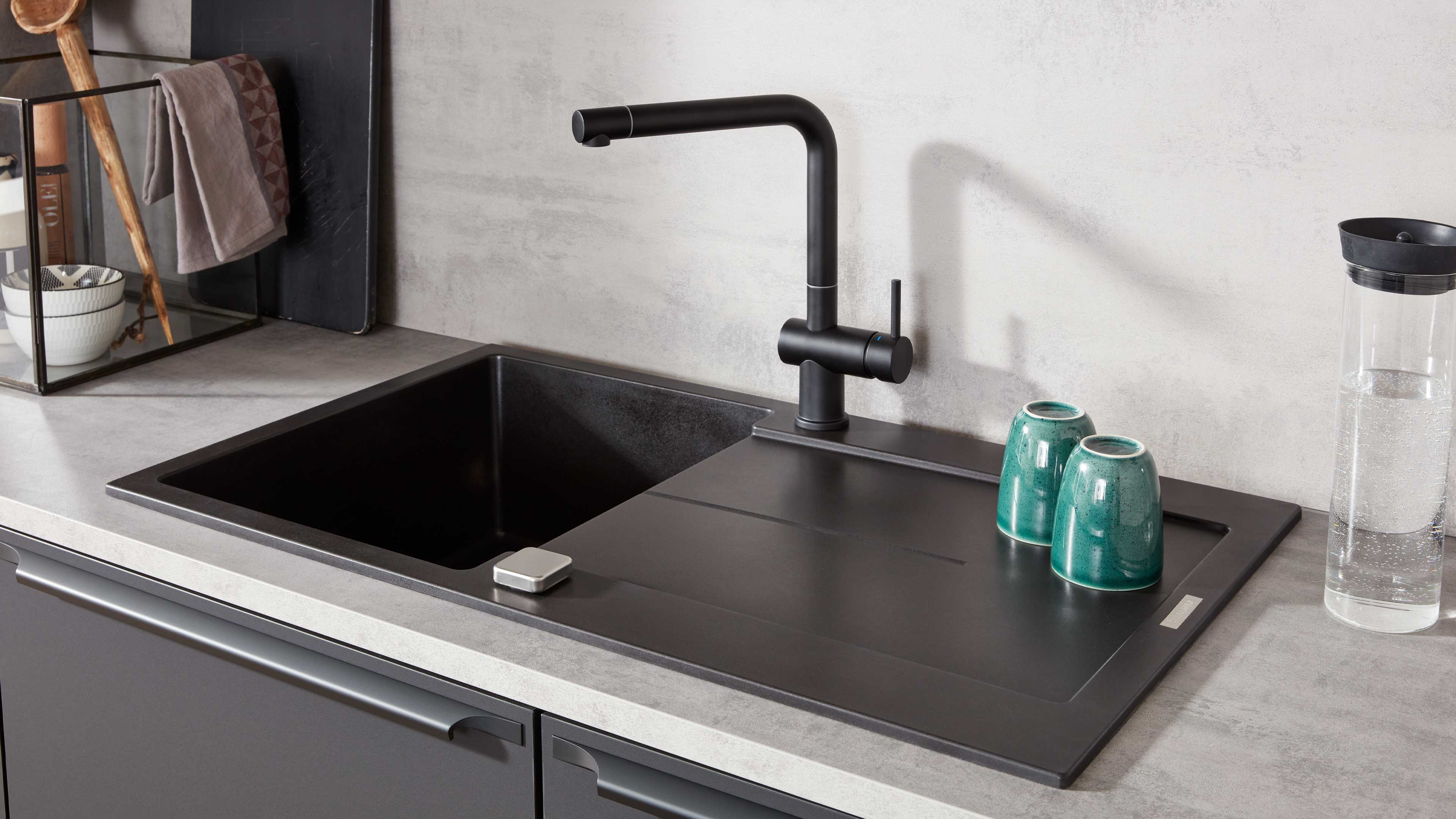 Schwarze Einbauspüle mit Abtropffläche, matt-schwarzer Armatur, zwei grüne Tassen und transparente Trinkflasche rechts.