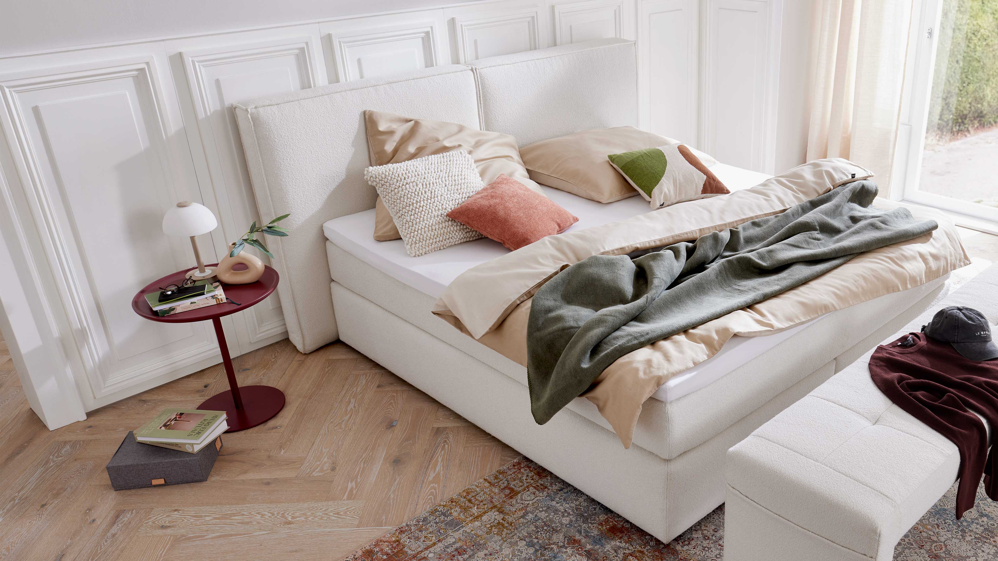 Weißes Doppelbett mit geteiltem Polsterkopfteil, beiger Bettwäsche, mehreren Kissen und grünem Plaid sowie rotem Beistelltisch mit Lampe.