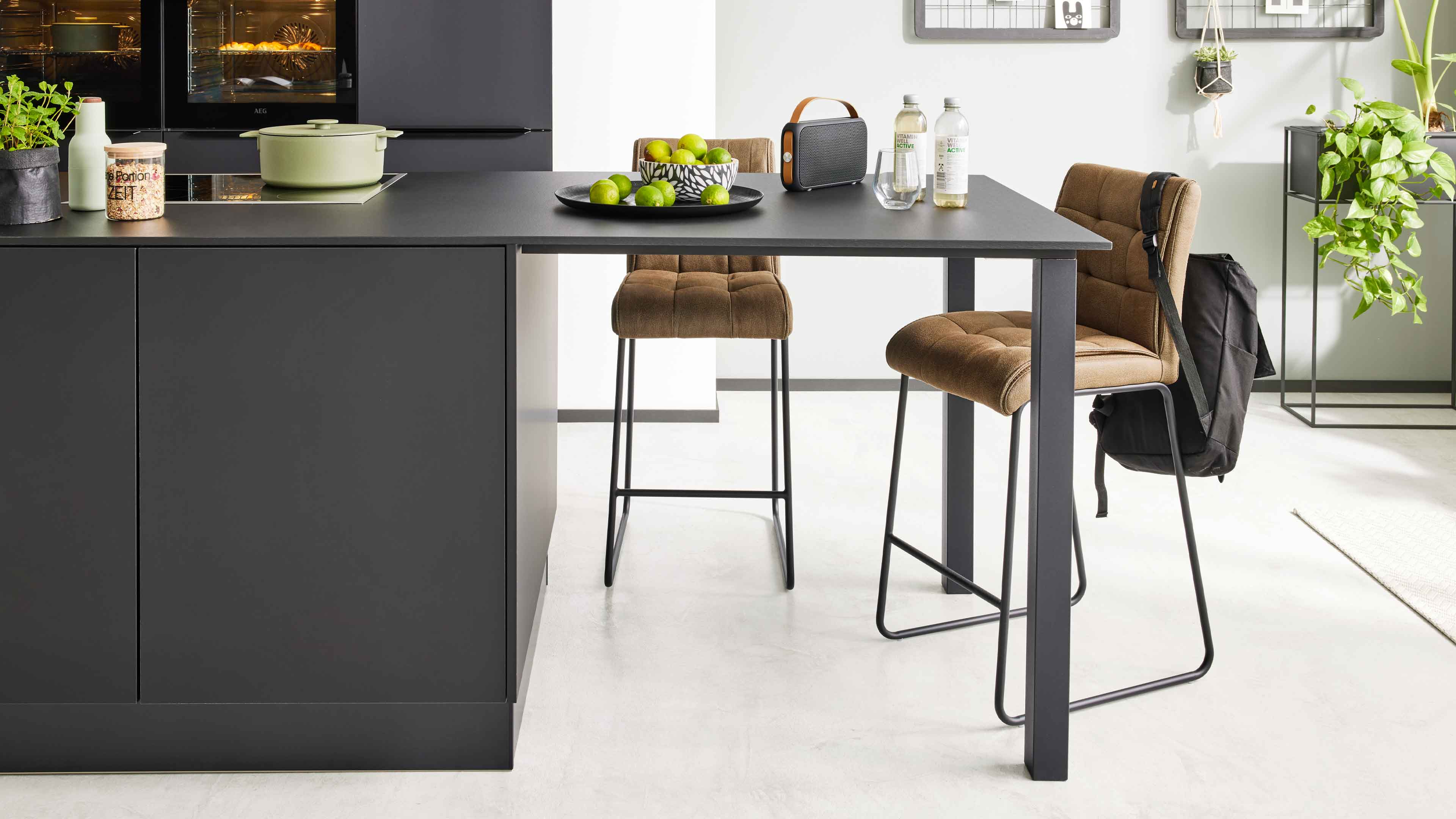 Schwarze Kücheninsel mit Bartisch, zwei braune gesteppte Barhocker auf Metallgestellen und an einem Hocker hängendem schwarzem Rucksack.