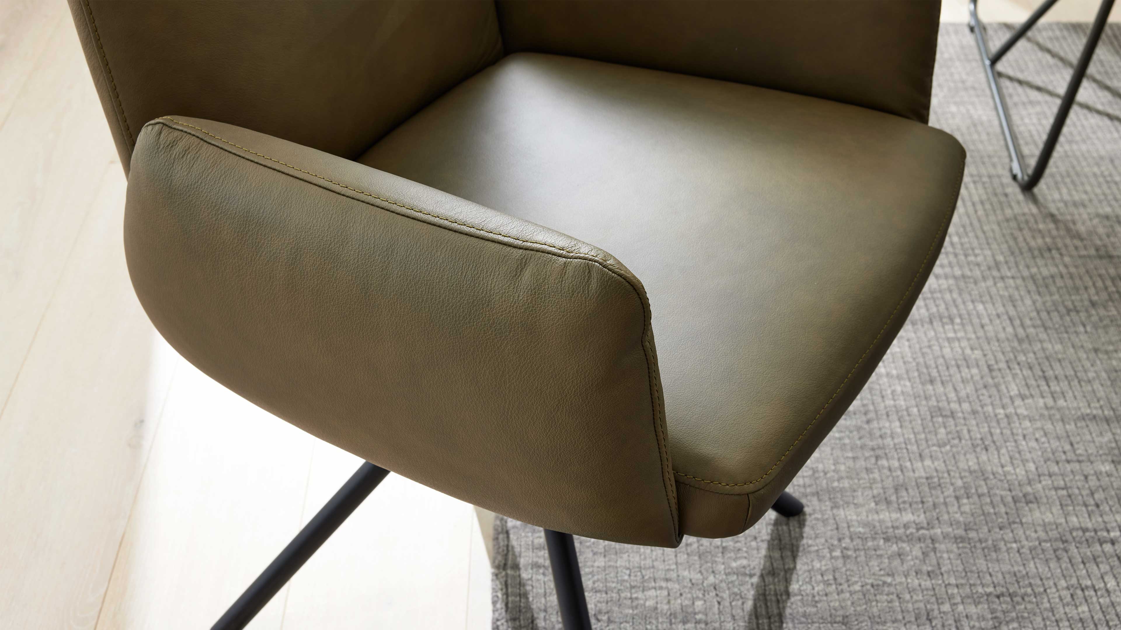 Nahaufnahme eines olivgrünen Ledersessels mit sichtbarer Naht, schwarzem Metallfuß und grauem Teppich.