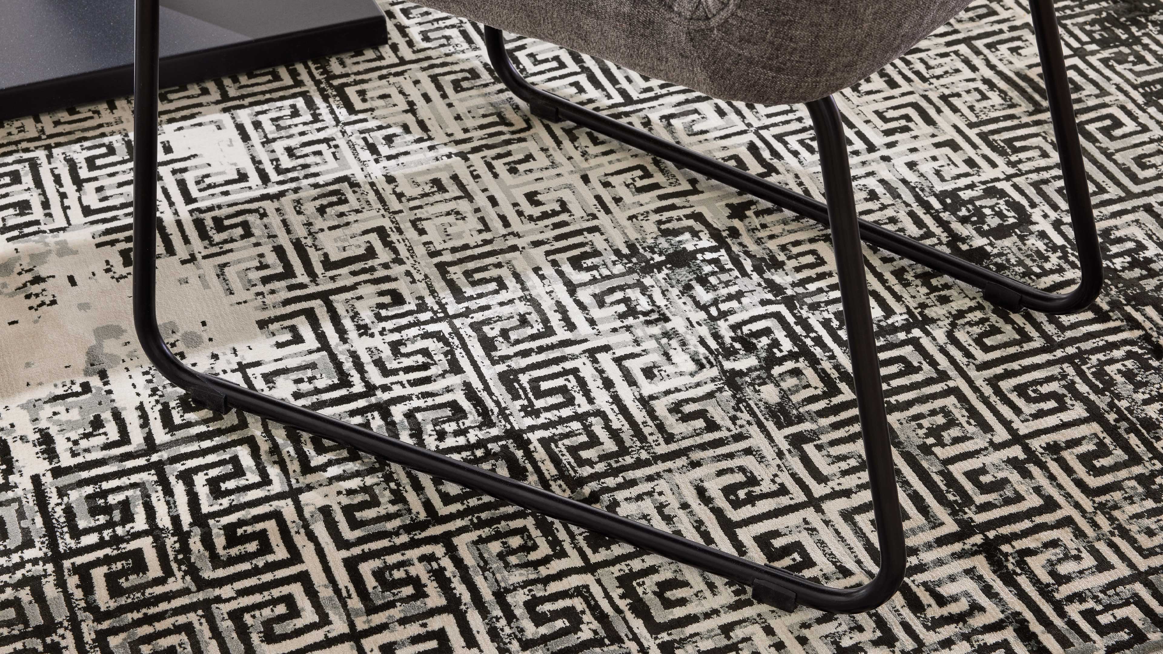 Schwarzes Metallgestell eines Stuhluntergestells mit grauem Polstersitz, auf einem beige-schwarzen Teppich mit geometrischem Labyrinthmuster.