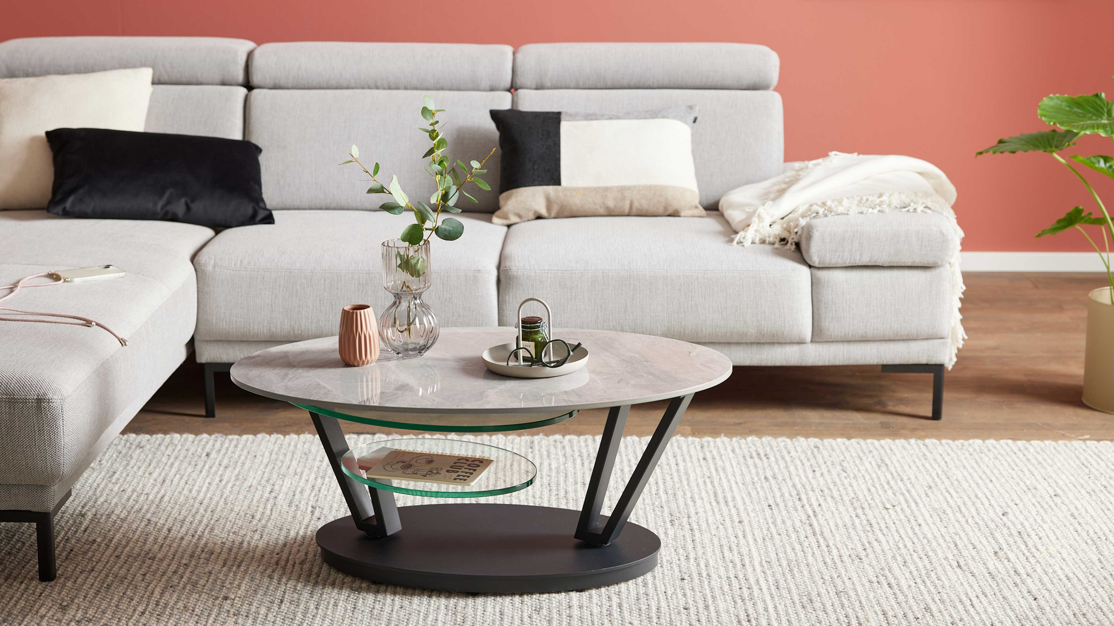 Runder Couchtisch mit heller Marmorplatte und Glasvase mit Eukalyptuszweigen, im Hintergrund hellgraues Ecksofa mit Kissen und Decke.